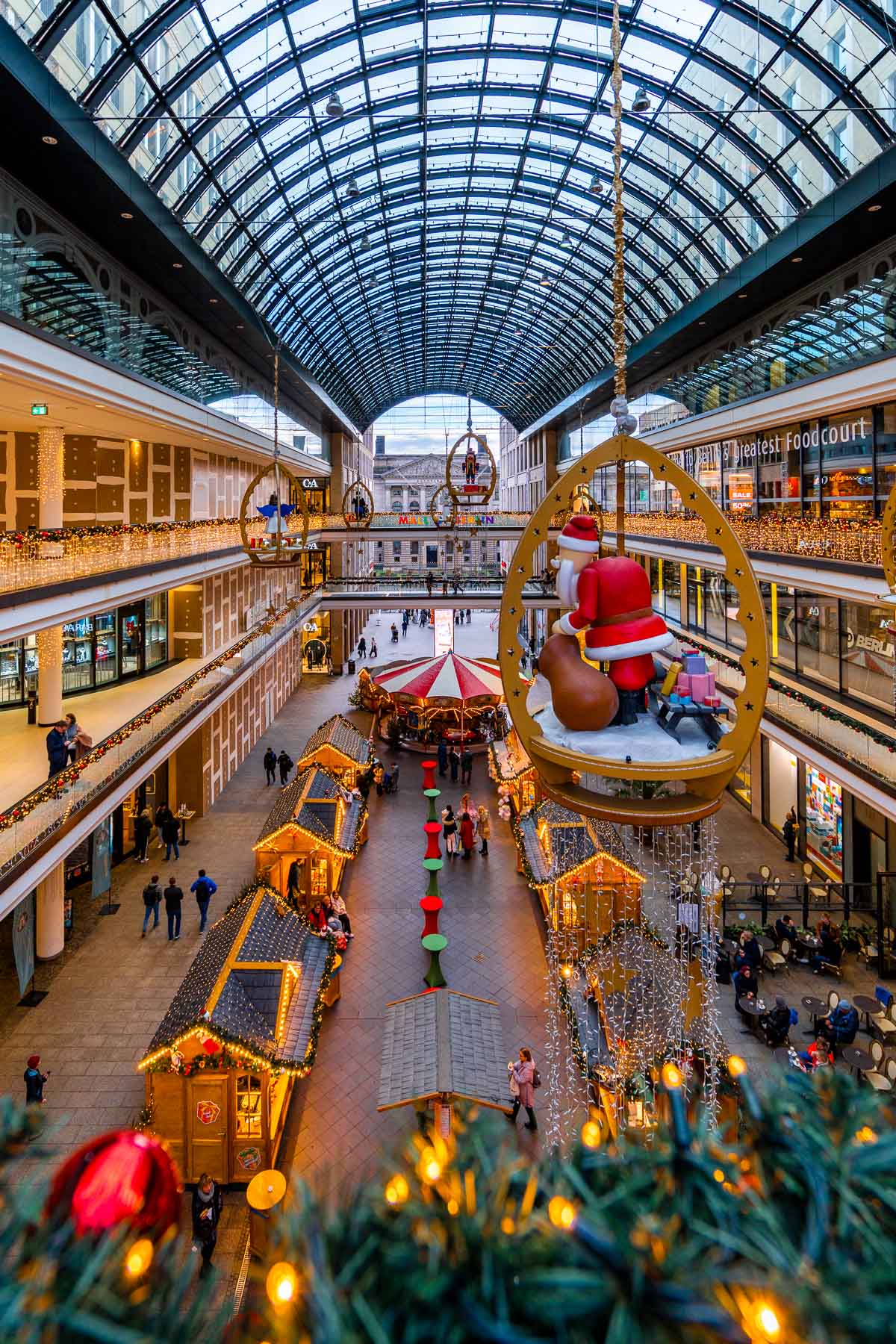 An indoor Christmas market fills the atrium of the Mall of Berlin, with wooden stalls, hanging Santa decorations, a carousel, and shoppers walking between the displays under a glass roof.