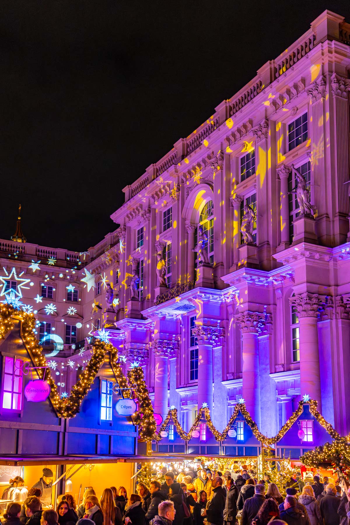 The Christmas market at the Humboldt Forum in Berlin with illuminated stalls, garlands, and a large historic building lit in purple and gold behind the crowd.