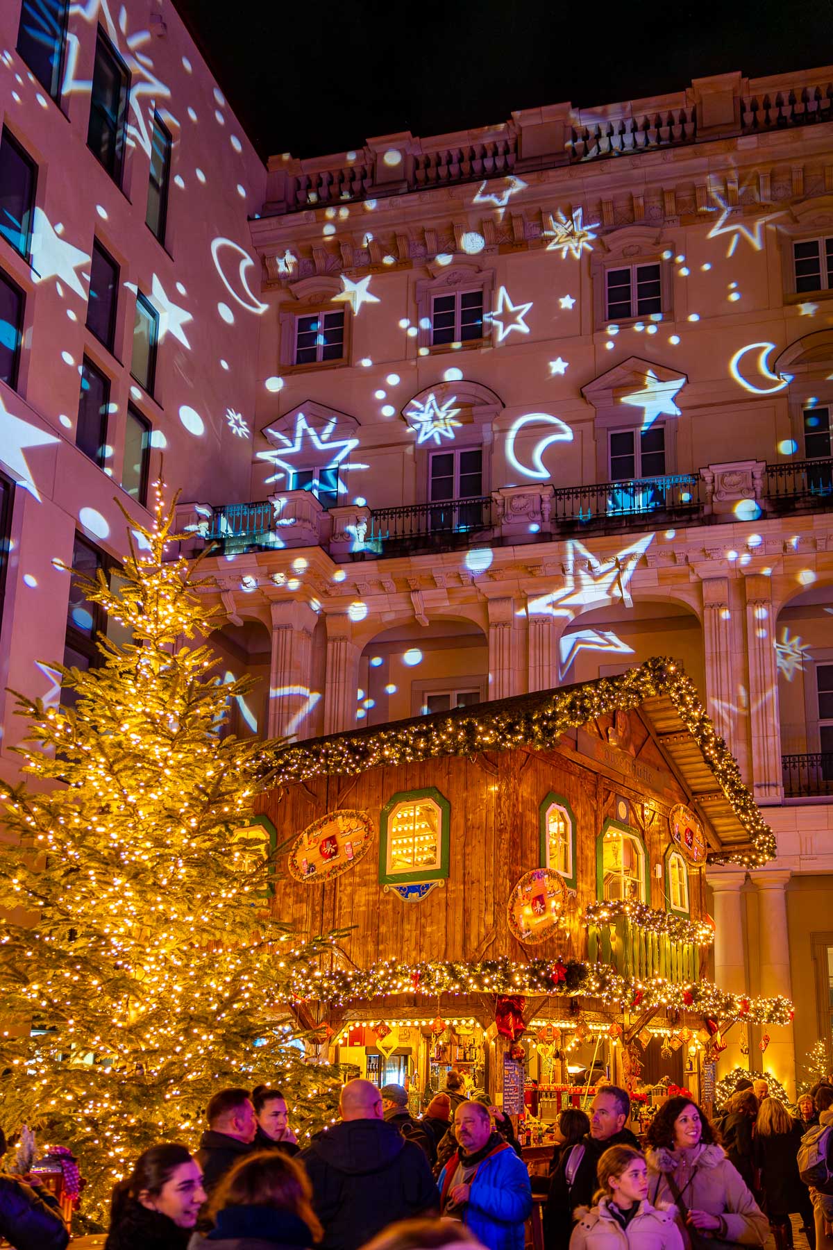 A wooden Christmas market house at the Humboldt Forum in Berlin decorated with lights and greenery, set in front of a building covered with projected stars and moon shapes.