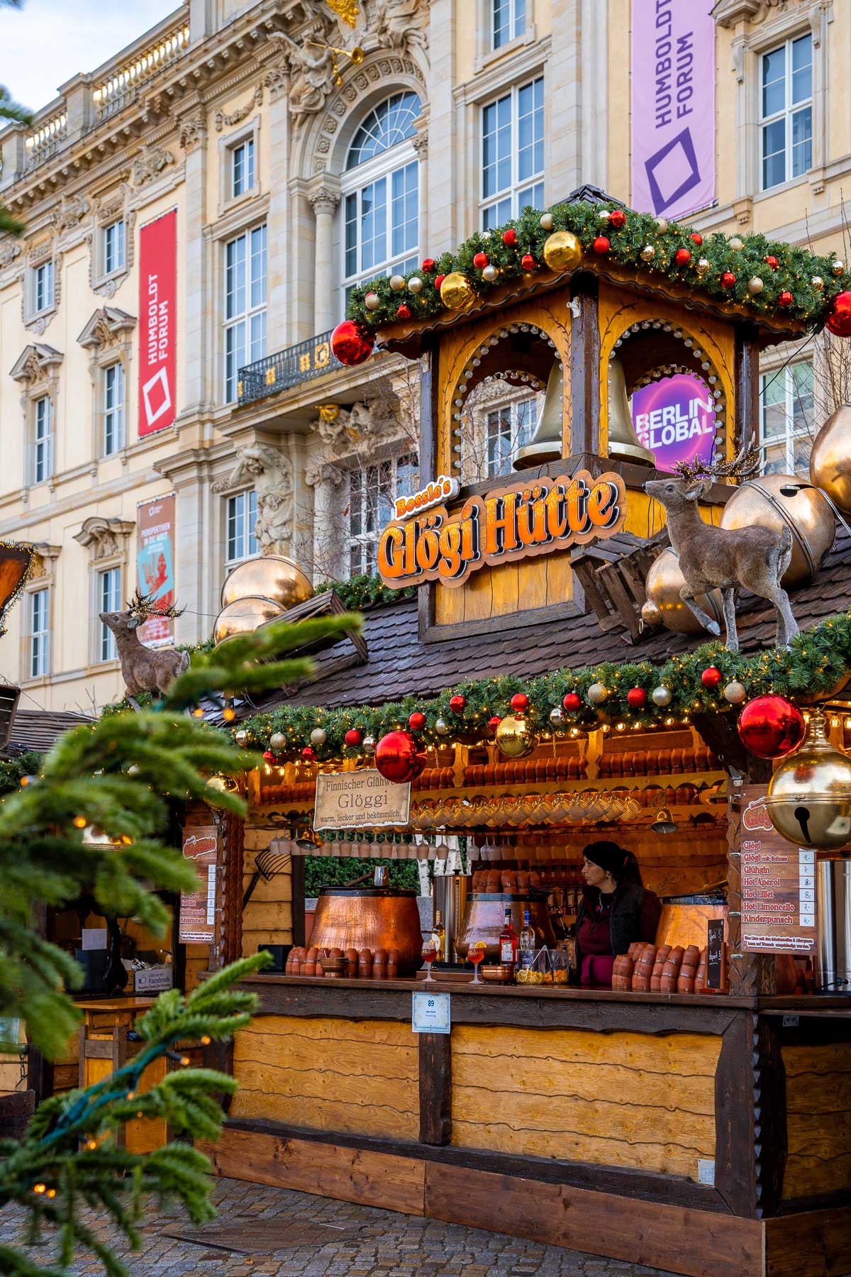 A wooden Glögi stand with garlands, ornaments, and reindeer figures sits in front of the Humboldt Forum building at the Christmas market at the Humboldt Forum, Berlin.