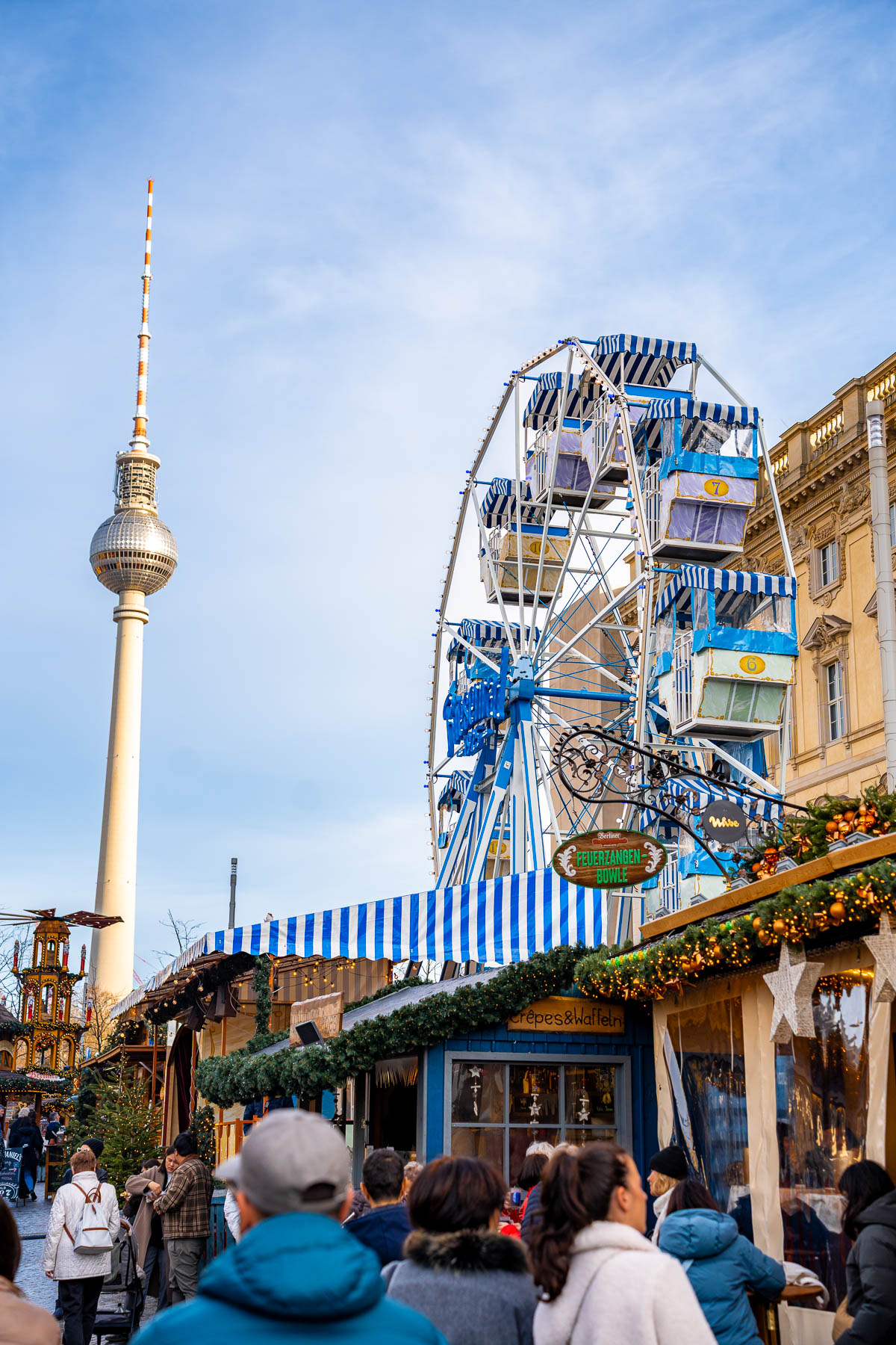 A Ferris wheel with blue and white gondolas stands behind Christmas market stalls, with the Berlin TV Tower visible in the background and people walking between decorated booths.