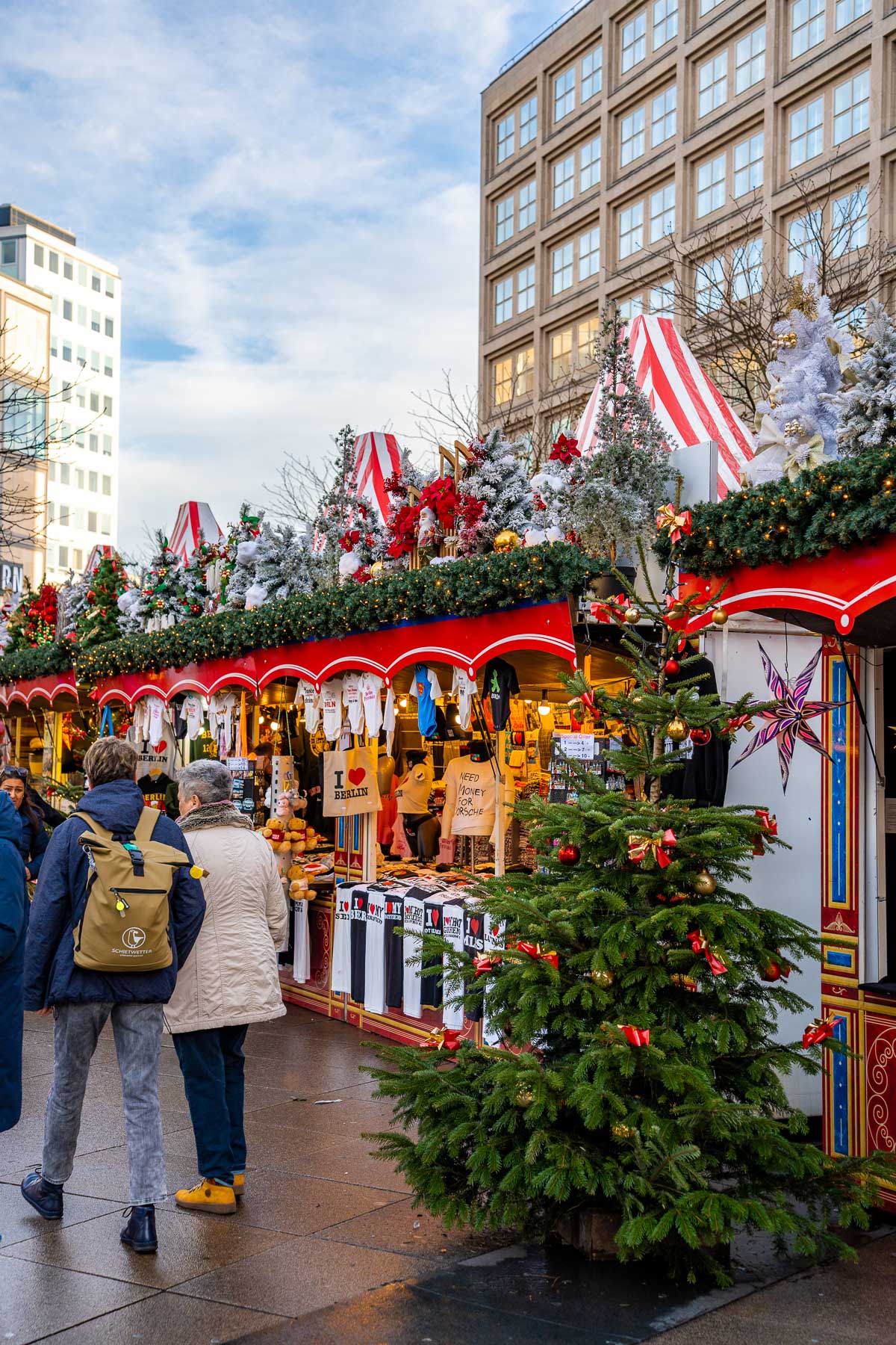 Rows of red and white striped market stalls line Alexanderplatz, with Christmas trees, hanging decorations, and shoppers browsing souvenirs and gifts.