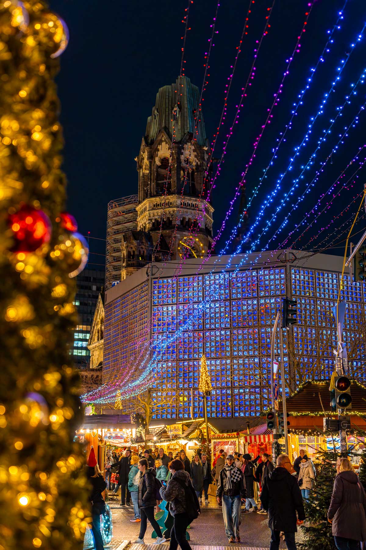 A night view of the Christmas Market on Breitscheidplatz, Berlin with the Kaiser Wilhelm Memorial Church in the background, colorful light strands overhead, market stalls, and people walking through the square.