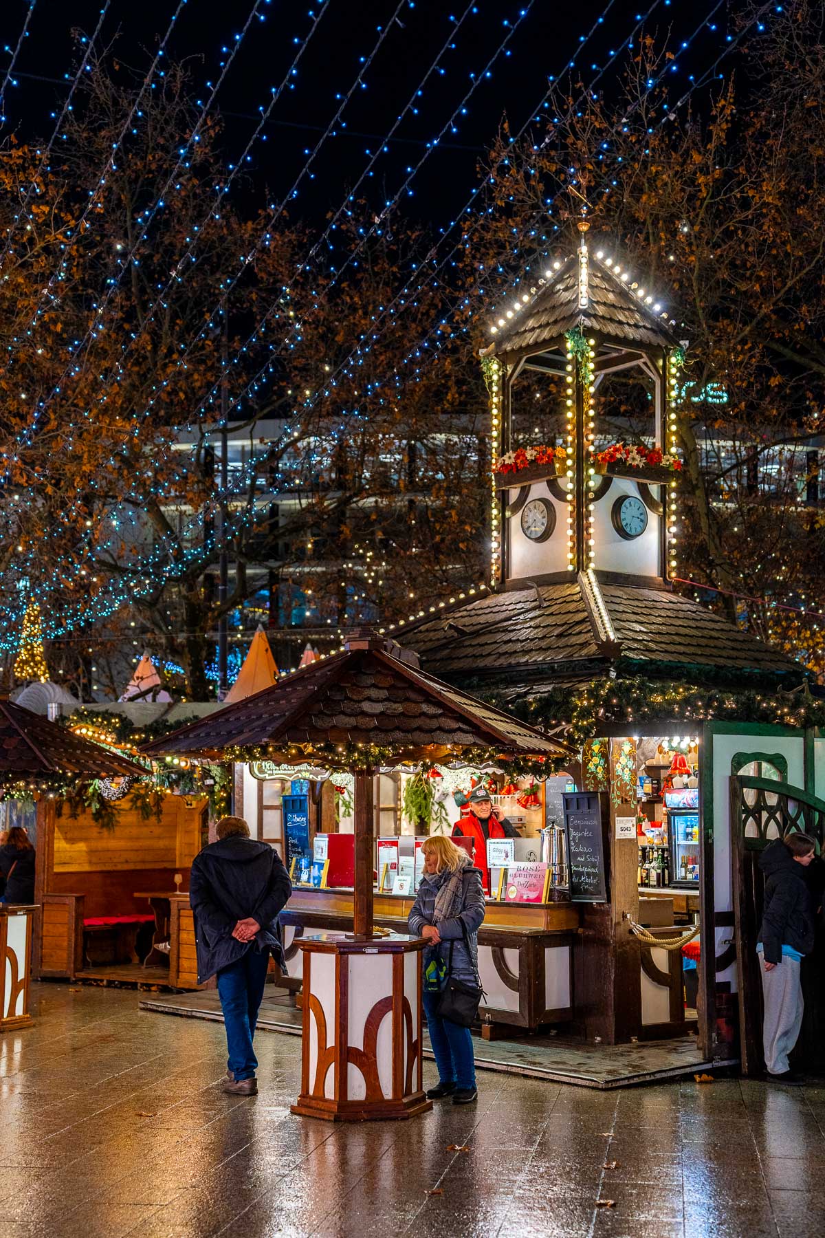 A wooden Christmas market stall at the Christmas Market on Breitscheidplatz, Berlin with a small clock tower roof, string lights, surrounding trees, and people standing nearby.
