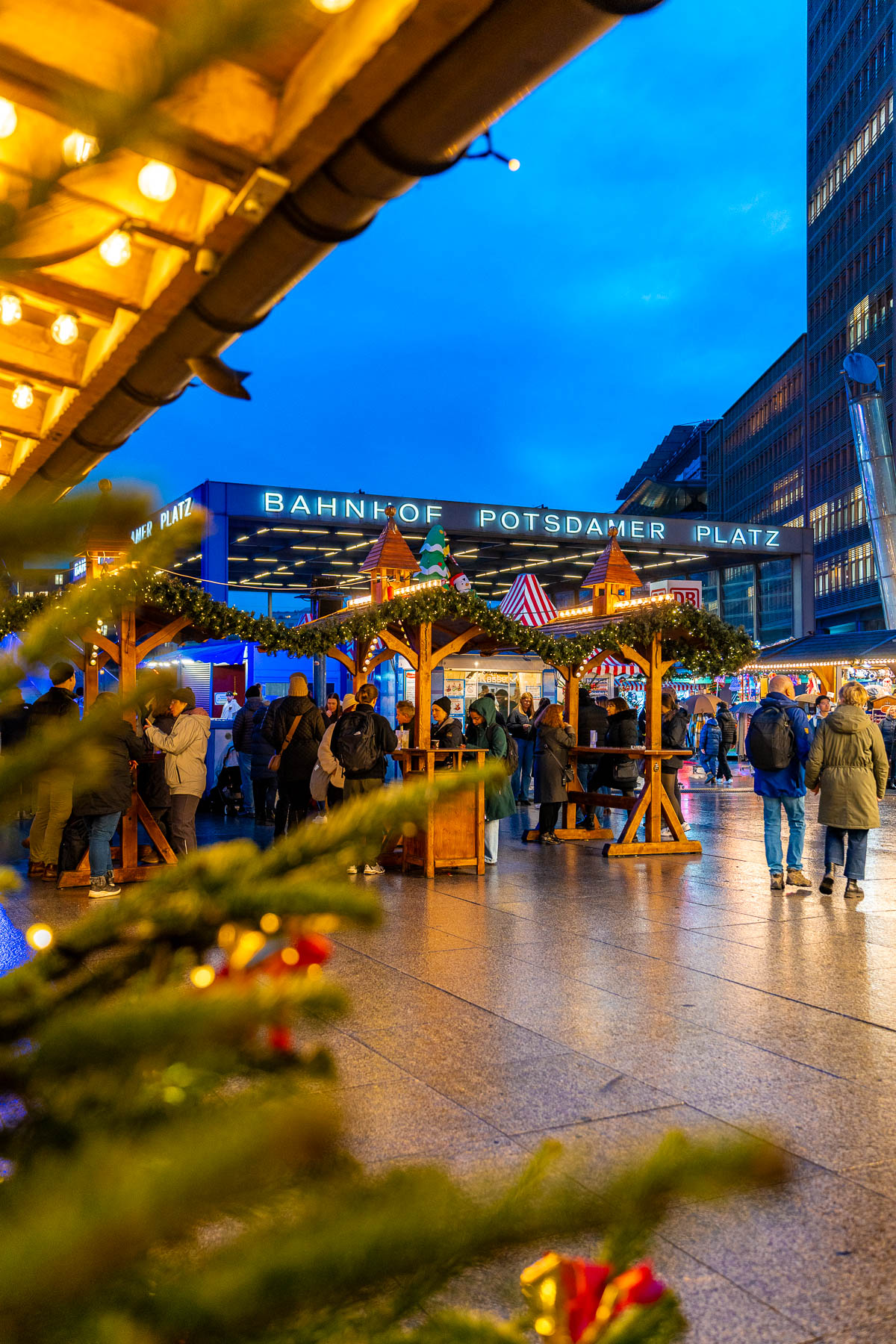 The Christmas Market on Potsdamer Platz, Berlin with wooden huts, standing tables, groups of people, and the Bahnhof Potsdamer Platz sign visible in the background at night.