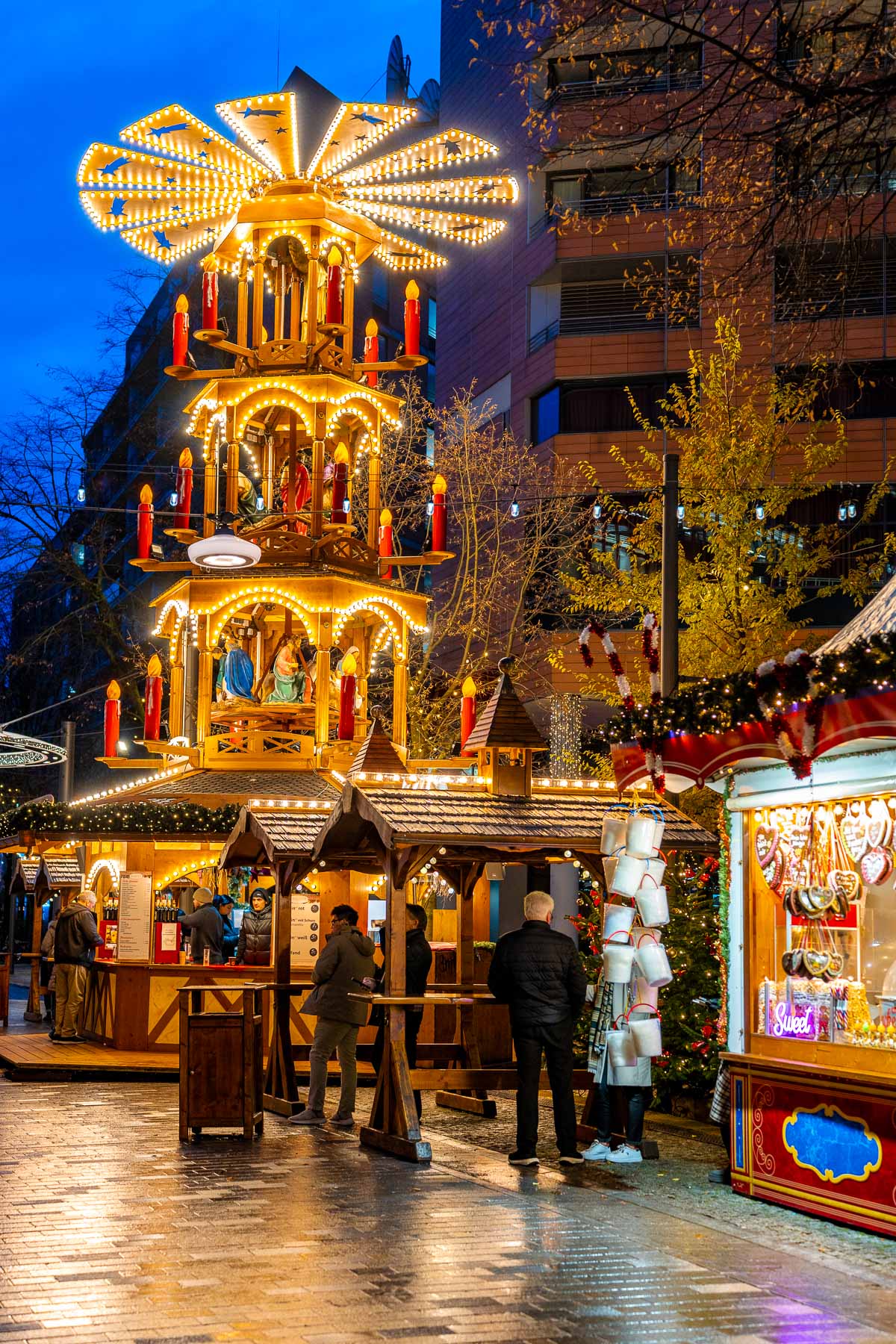 A wooden Christmas pyramid lit with warm bulbs stands at the Christmas Market on Potsdamer Platz, Berlin, with food stalls around it and people gathered nearby.