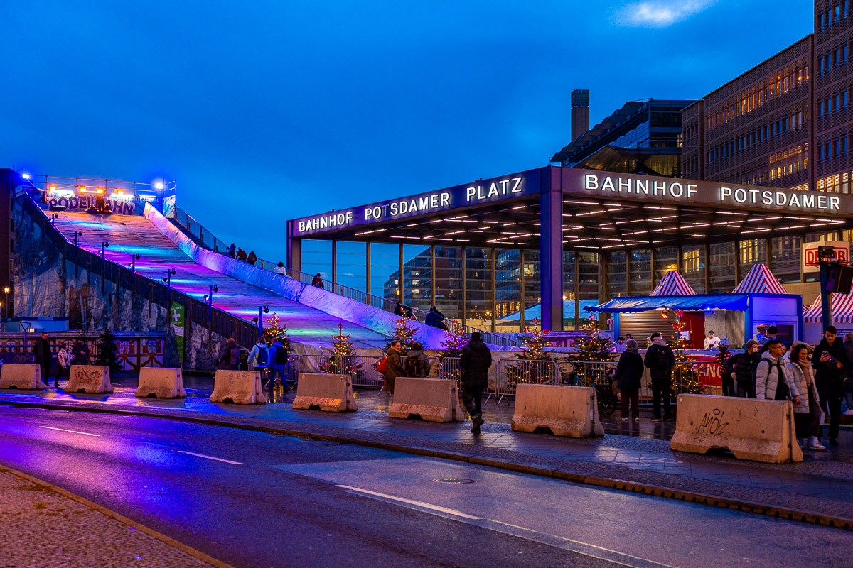 A wide view of the Christmas Market on Potsdamer Platz, Berlin with the Bahnhof Potsdamer Platz building, a lit sledding ramp, market stalls, small Christmas trees, and people walking in the evening.