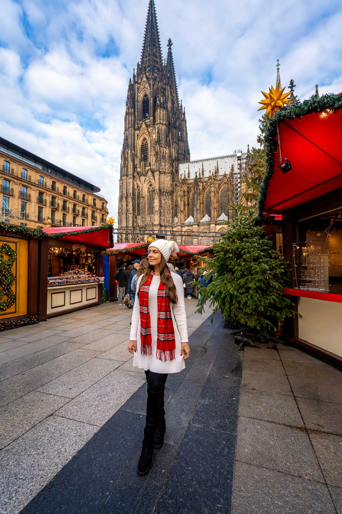 A woman walking between red Christmas market stalls with Cologne Cathedral rising in the background.