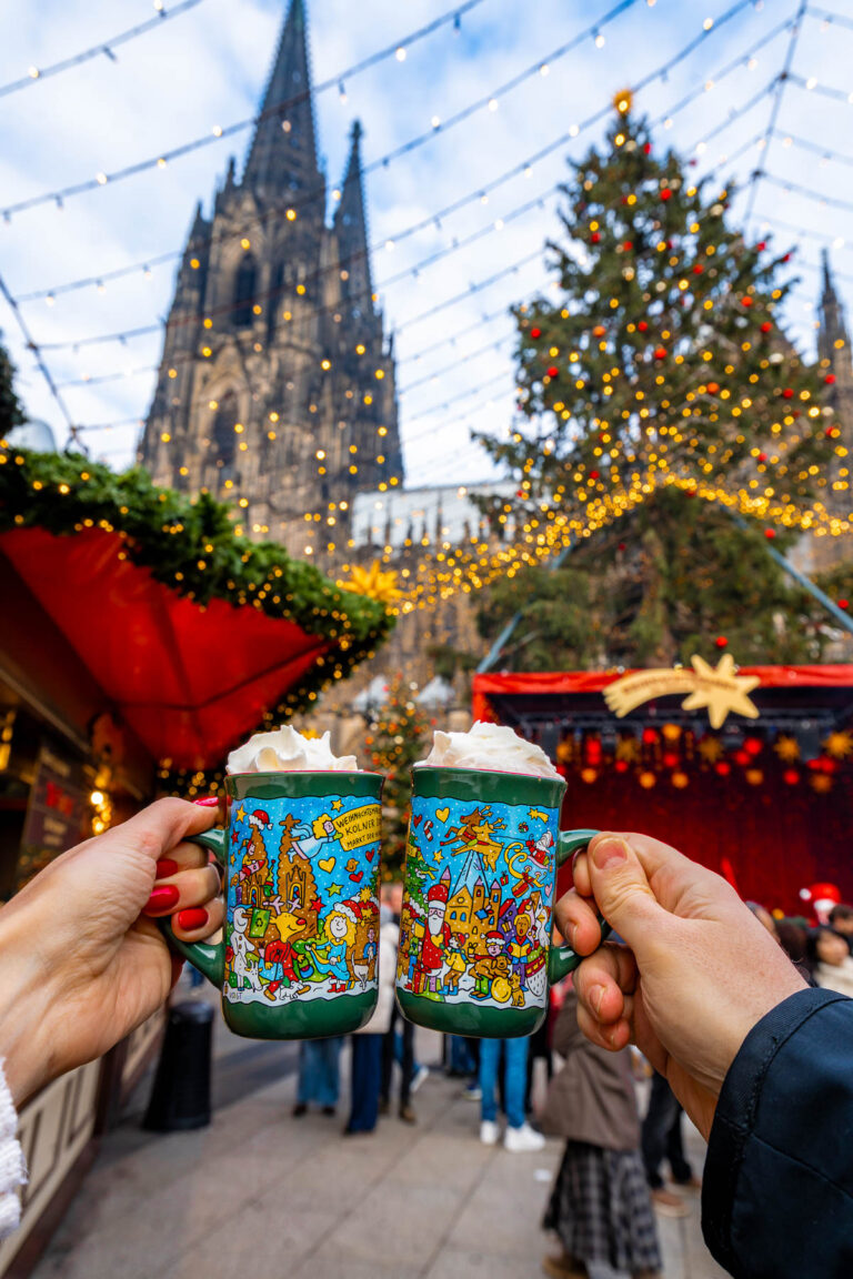 Two hands holding decorated mugs of hot drinks with Cologne Cathedral and Christmas lights behind them.