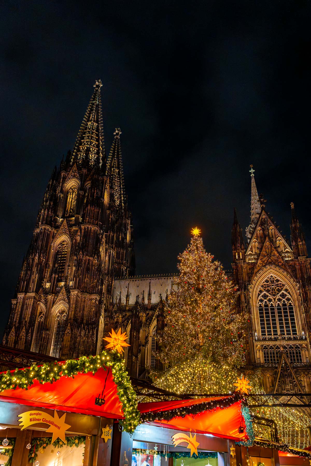 Night view of Cologne Cathedral with the Christmas market stalls in front and a large lit Christmas tree in the center.