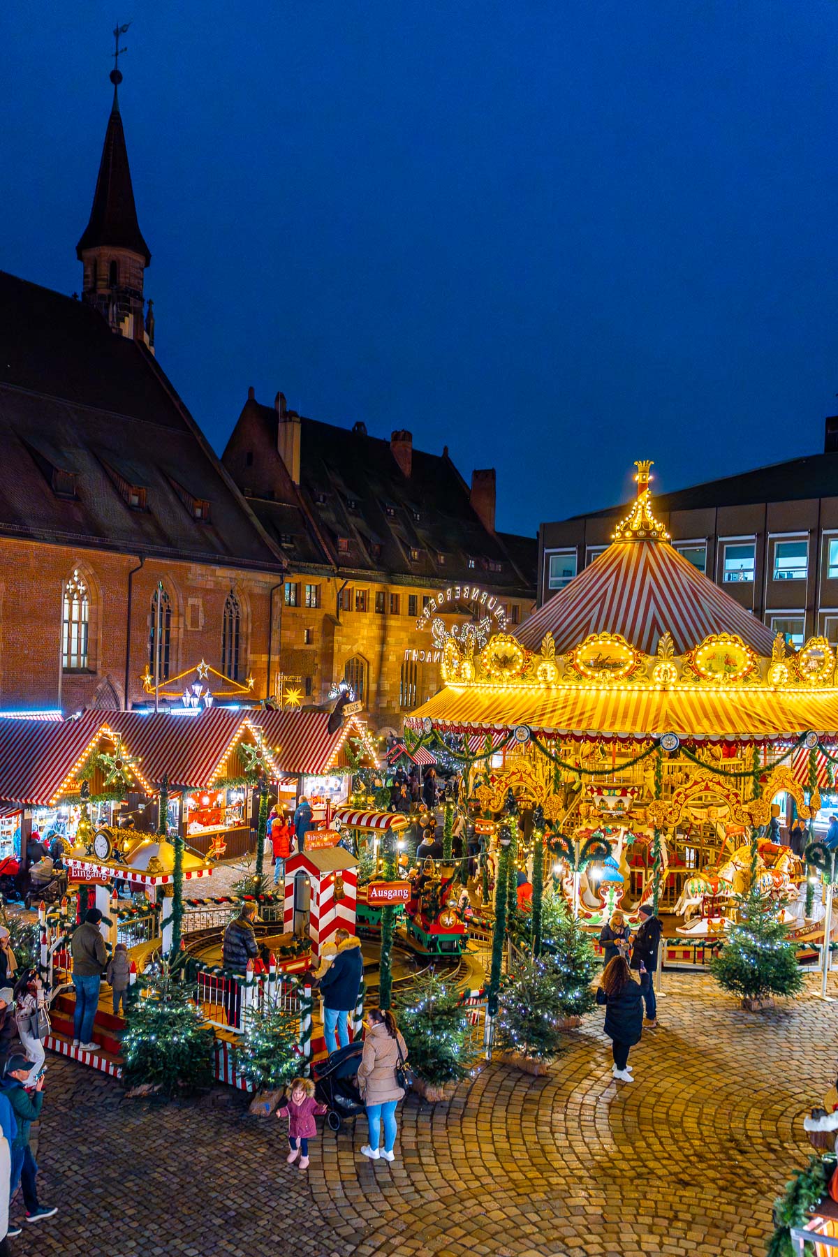 An elevated view of Kinderweihnacht Christkindlesmarkt in Nuremberg at night, showing a carousel, small decorated trees, striped stalls, and families walking through the square.