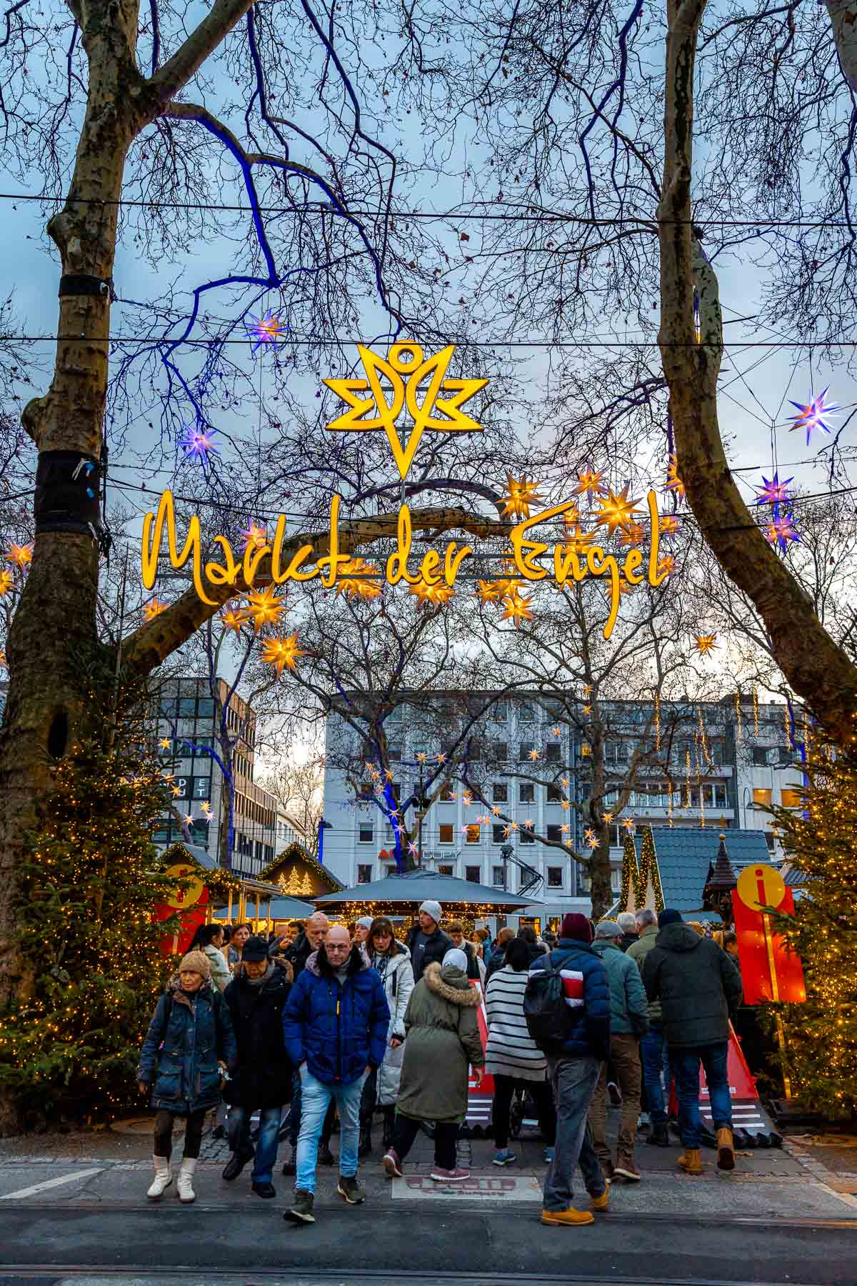 Entrance to the Market of Angels in Cologne with the illuminated sign hanging between trees and crowds walking underneath.