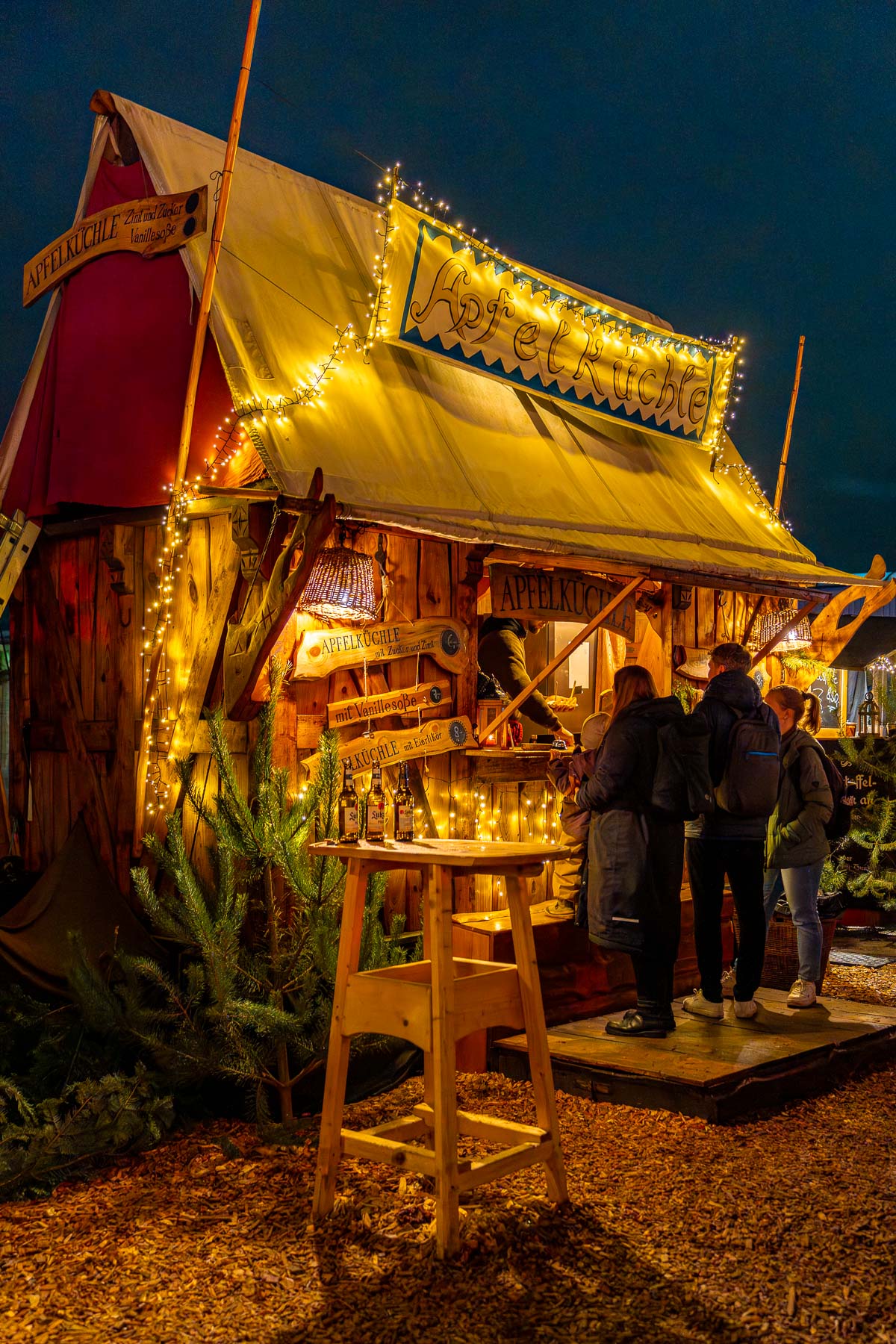 A small wooden food stall at the Medieval Christmas Market, Berlin, decorated with string lights, with people ordering food at the counter.
