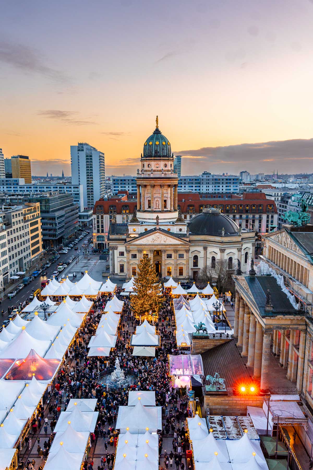 An aerial view of WeihnachtsZauber at Gendarmenmarkt in Berlin with white market tents, a large Christmas tree in the center, crowds filling the square, and the Deutscher Dom in the background at sunset.