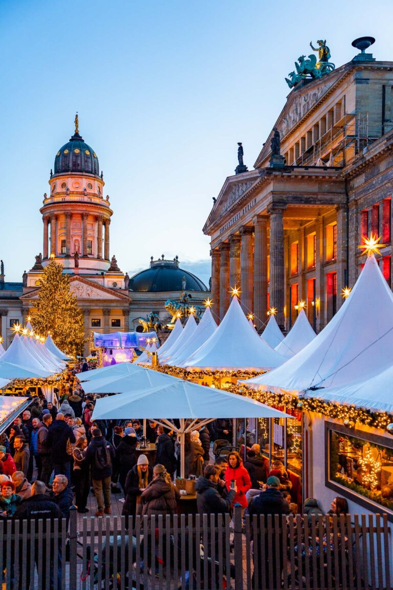 View of WeihnachtsZauber at Gendarmenmarkt in Berlin showing rows of white tents, people standing and walking between stalls, the Konzerthaus Berlin on the right, and the Deutscher Dom behind the Christmas tree.