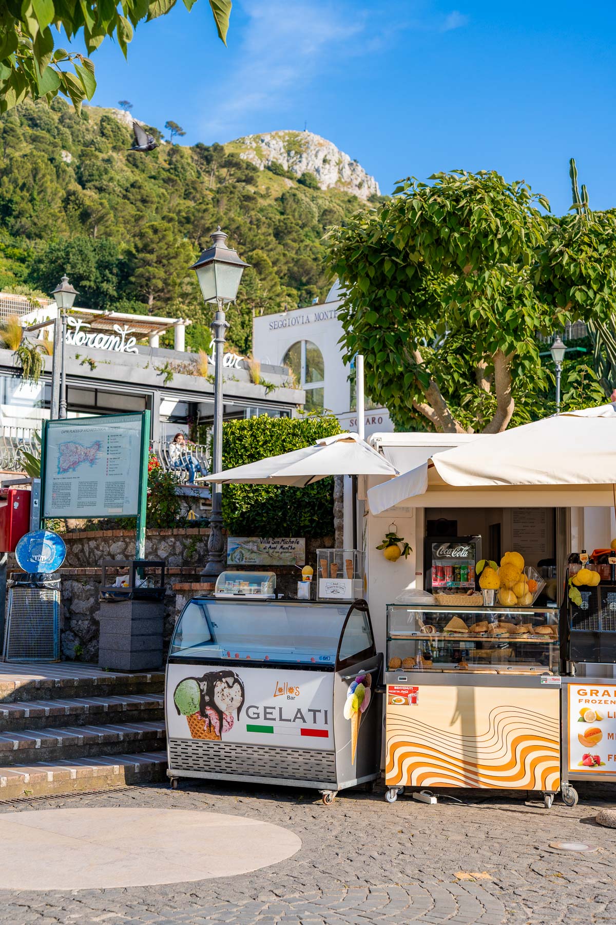Small food stand in Anacapri, Capri, Italy selling gelato and drinks, with display carts, umbrellas, lemon decorations, and green hills rising behind the square.