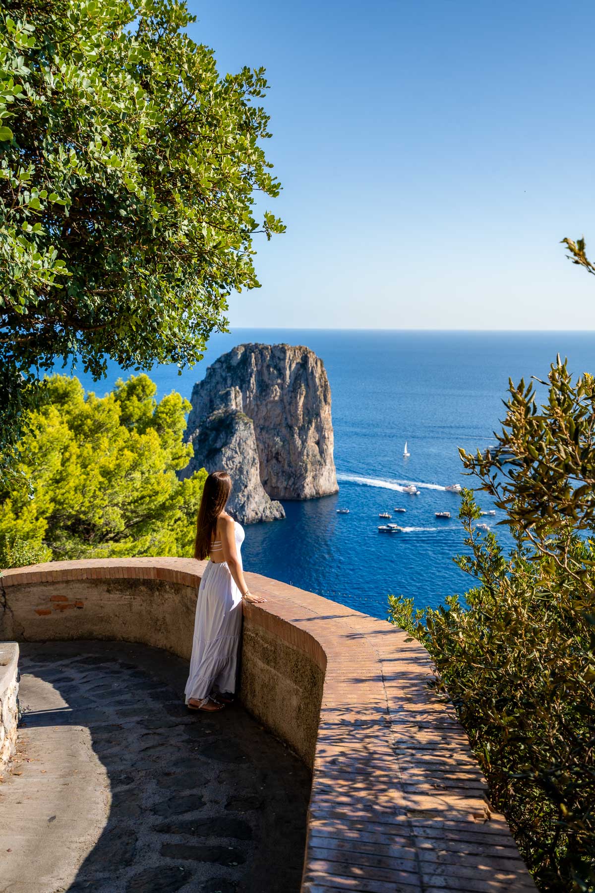 A woman standing at Belvedere Tragara in Capri, wearing a white dress and looking out over the Faraglioni rock, with stone paths, trees, and boats visible below.