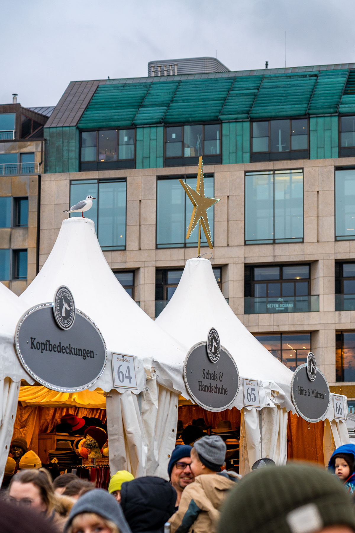 White tented stalls at the Christmas Market at Jungfernstieg, Hamburg, selling hats, scarves, and gloves, with crowds gathered in front and a modern building in the background.
