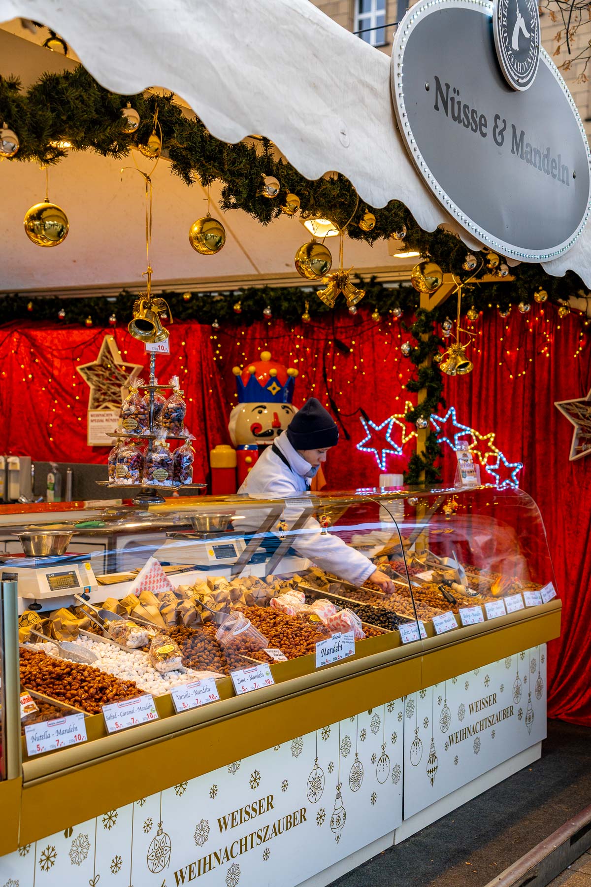 Vendor working behind a nuts and sweets stall at the Christmas Market at Jungfernstieg, Hamburg, with glass display cases, hanging decorations, and a red backdrop inside the booth.