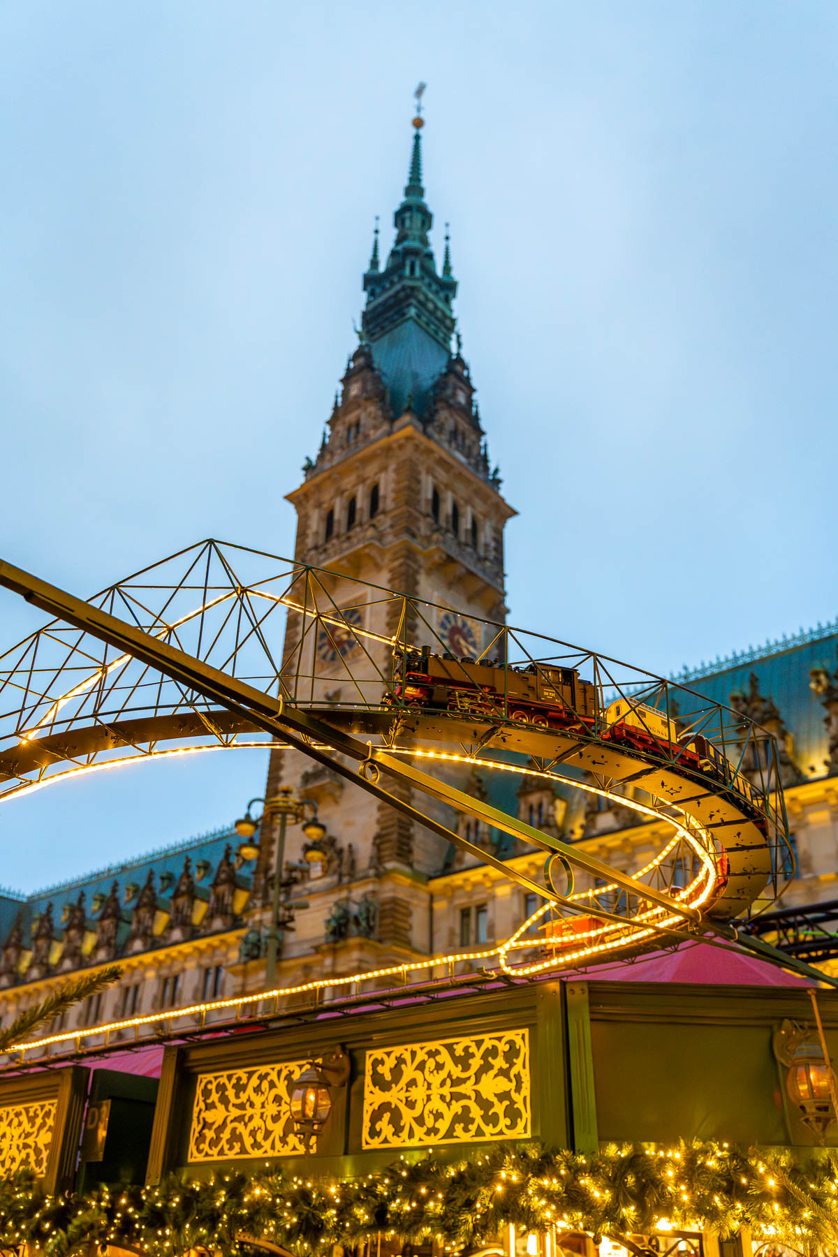 A small illuminated train ride running above the stalls at the Christmas Market at Rathausmarkt, Hamburg, with Hamburg City Hall rising in the background.