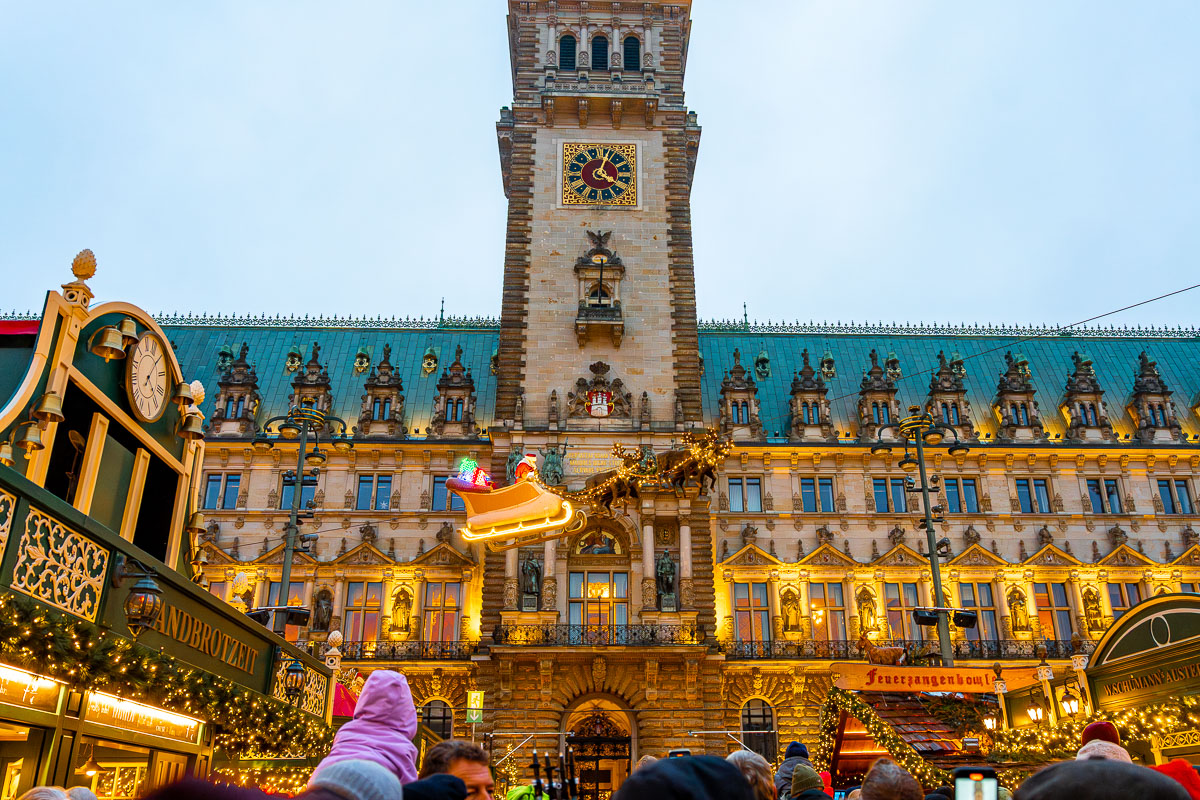Front view of Hamburg City Hall at Rathausmarkt with a Flying Santa in the middle, Christmas market stalls below, and a crowd gathered in the square.