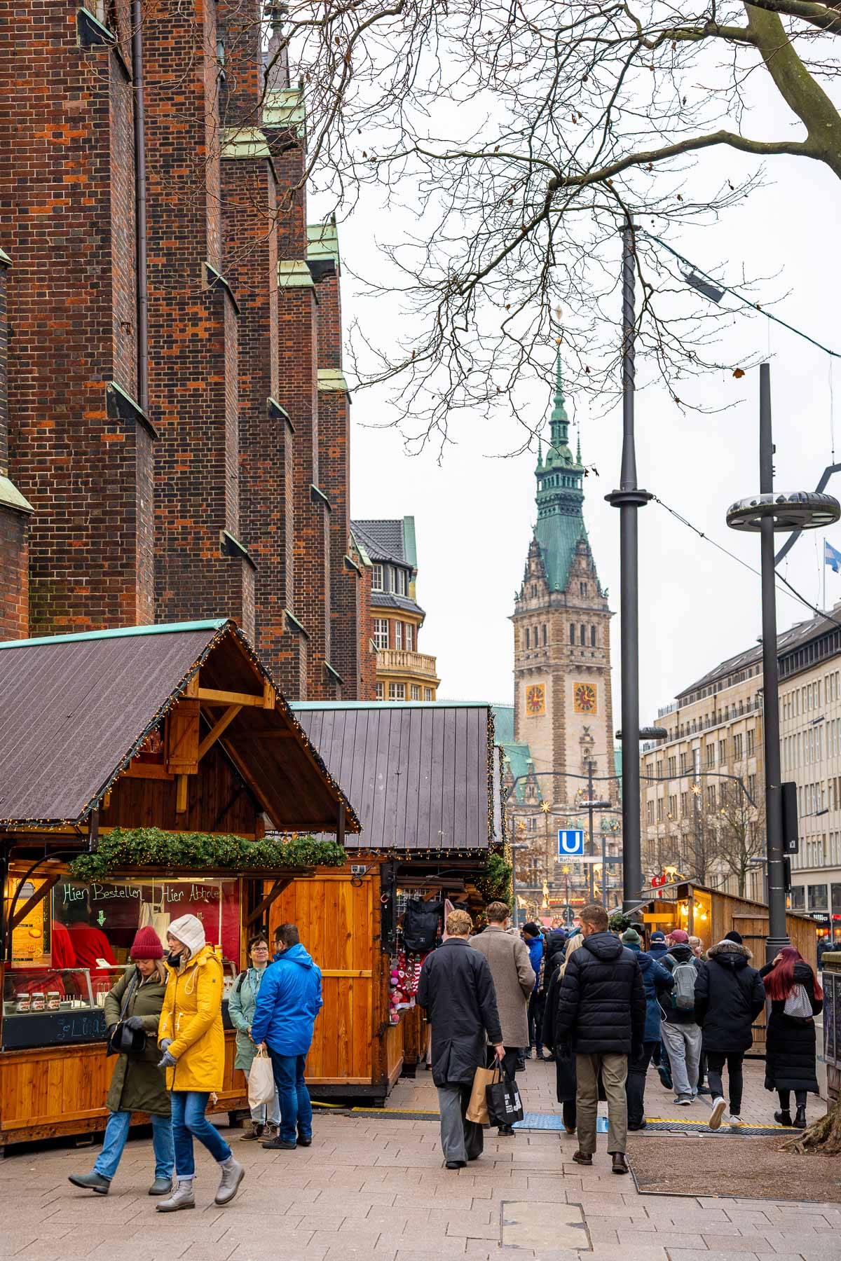 Daytime view of the Christmas market near St. Peter’s Church in Hamburg, with wooden stalls lining the street, people walking past, and the church tower visible in the background.