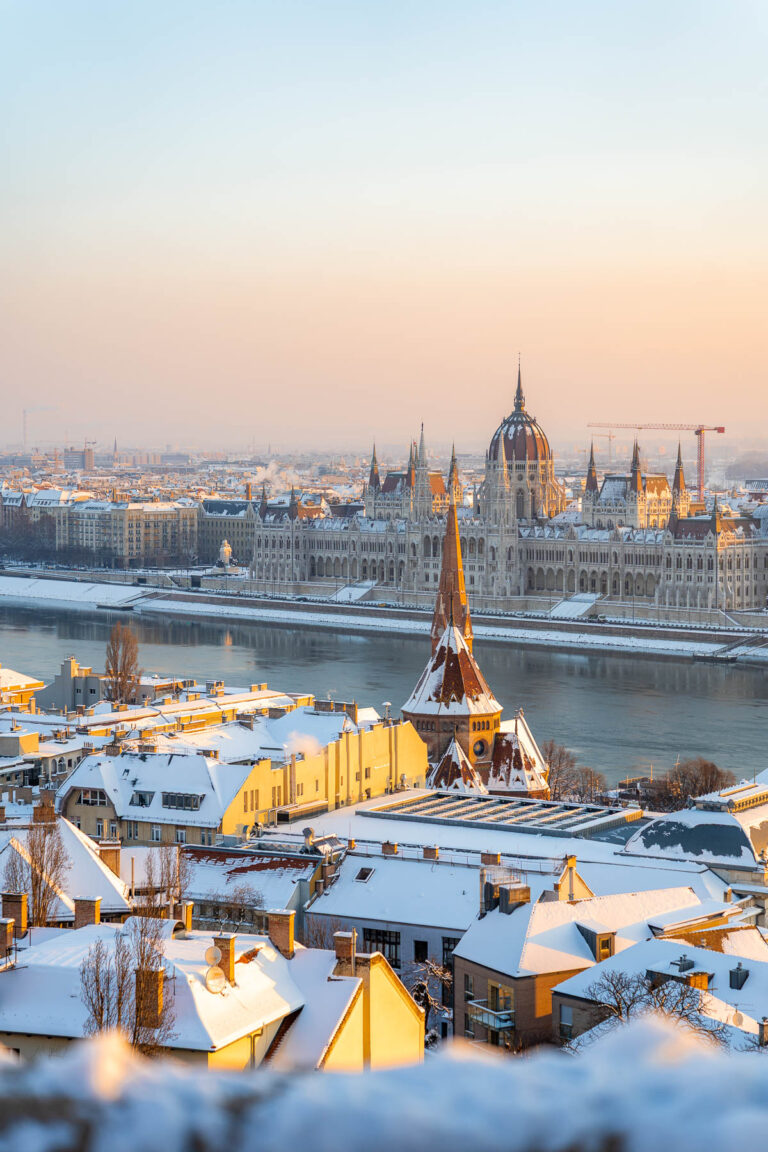 View from Fisherman’s Bastion in Budapest looking over snow covered rooftops, with the Danube River and the Hungarian Parliament building in the background.