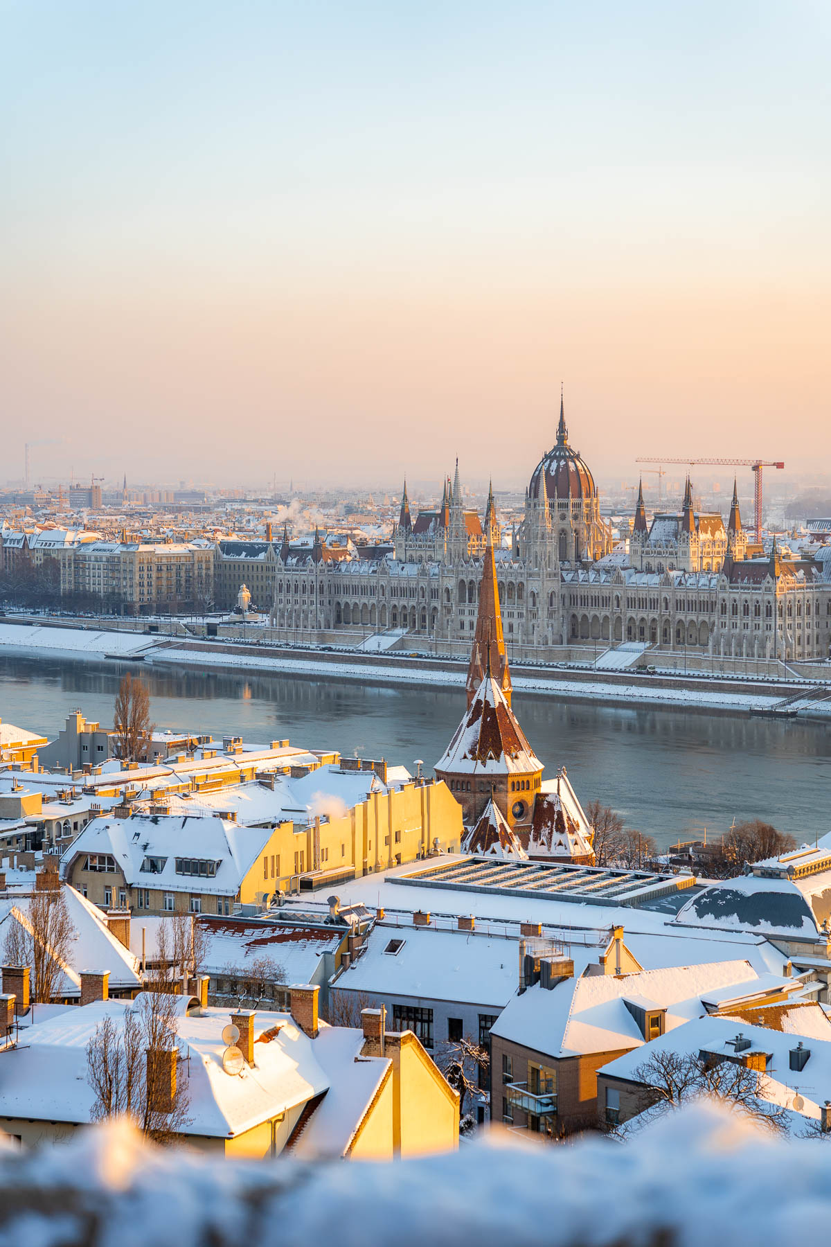 View from Fisherman’s Bastion in Budapest looking over snow covered rooftops, with the Danube River and the Hungarian Parliament building in the background.