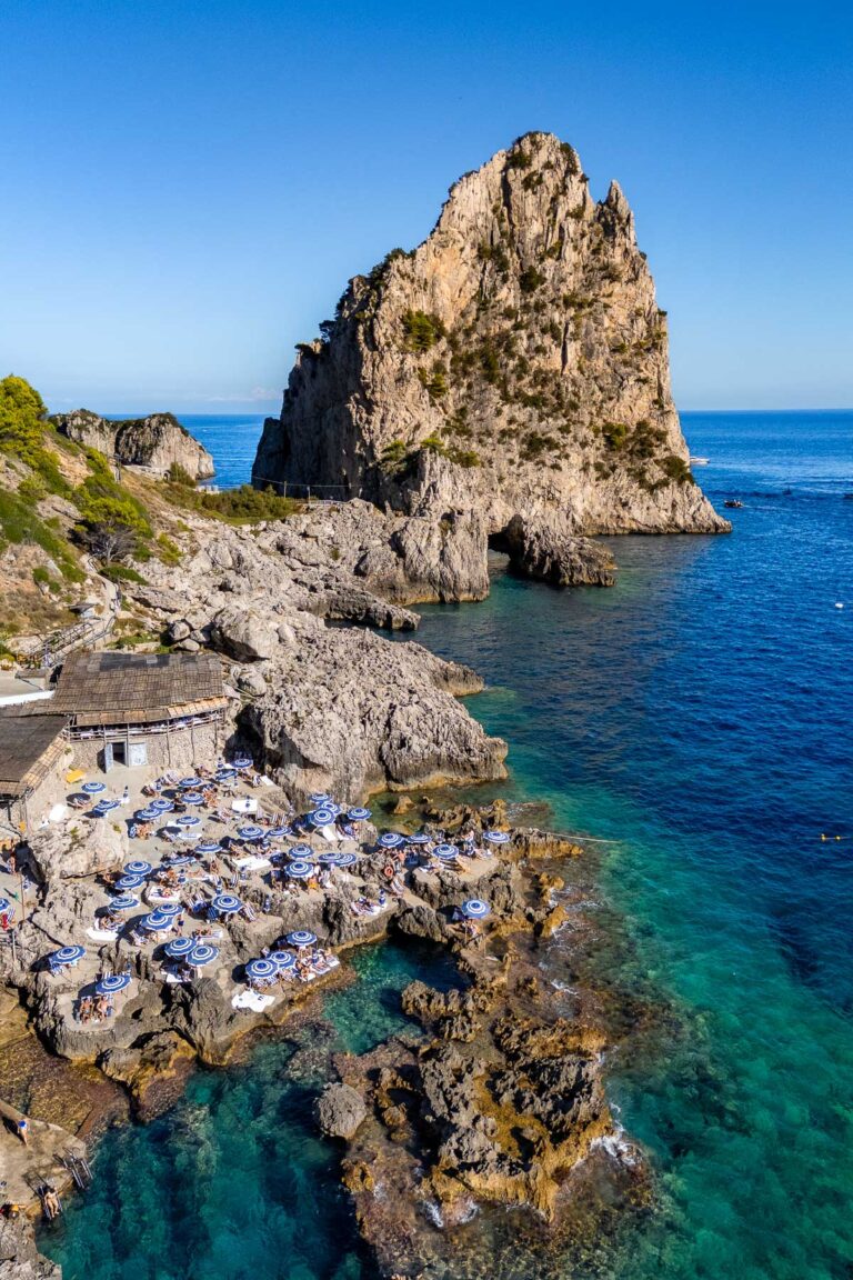Wide coastal view of La Fontelina Beach Club in Capri with rows of blue and white umbrellas on the rocks below, steep cliffs rising behind them, and deep blue water along the shoreline.