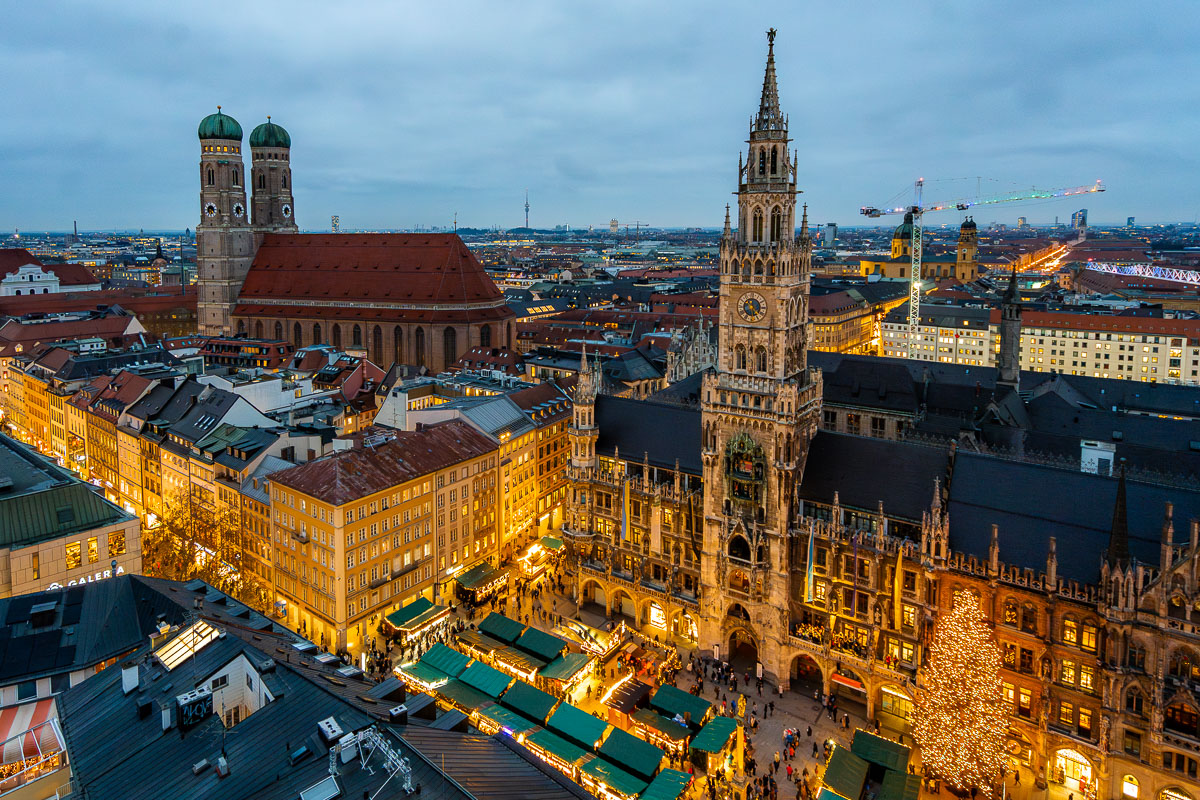 View of Marienplatz Christmas Market in Munich at dusk from St. Peter's Church, showing the New Town Hall tower, rows of green roofed market stalls, and crowds moving through the square.