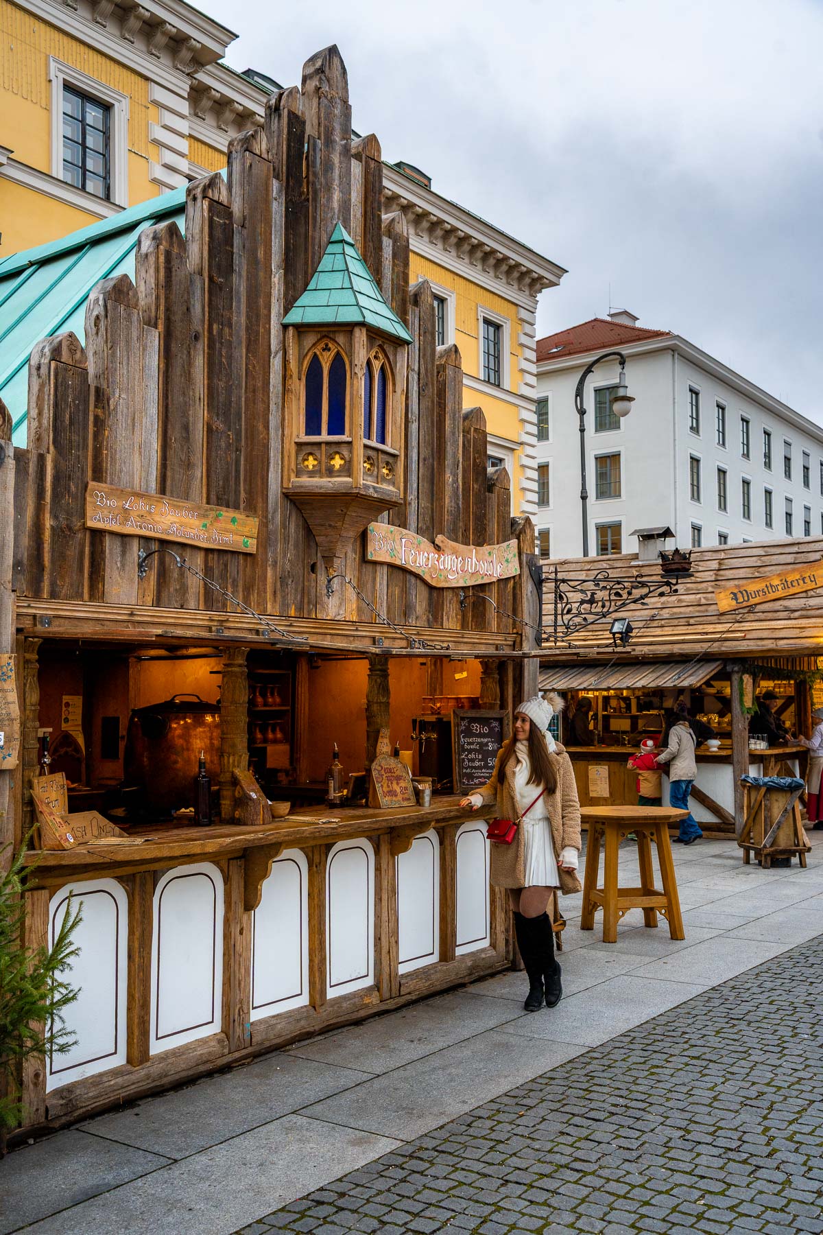A wooden stall at the Medieval Christmas Market in Munich with a carved facade and small tower detail, with a woman standing at the counter and other market stalls lined up behind her.