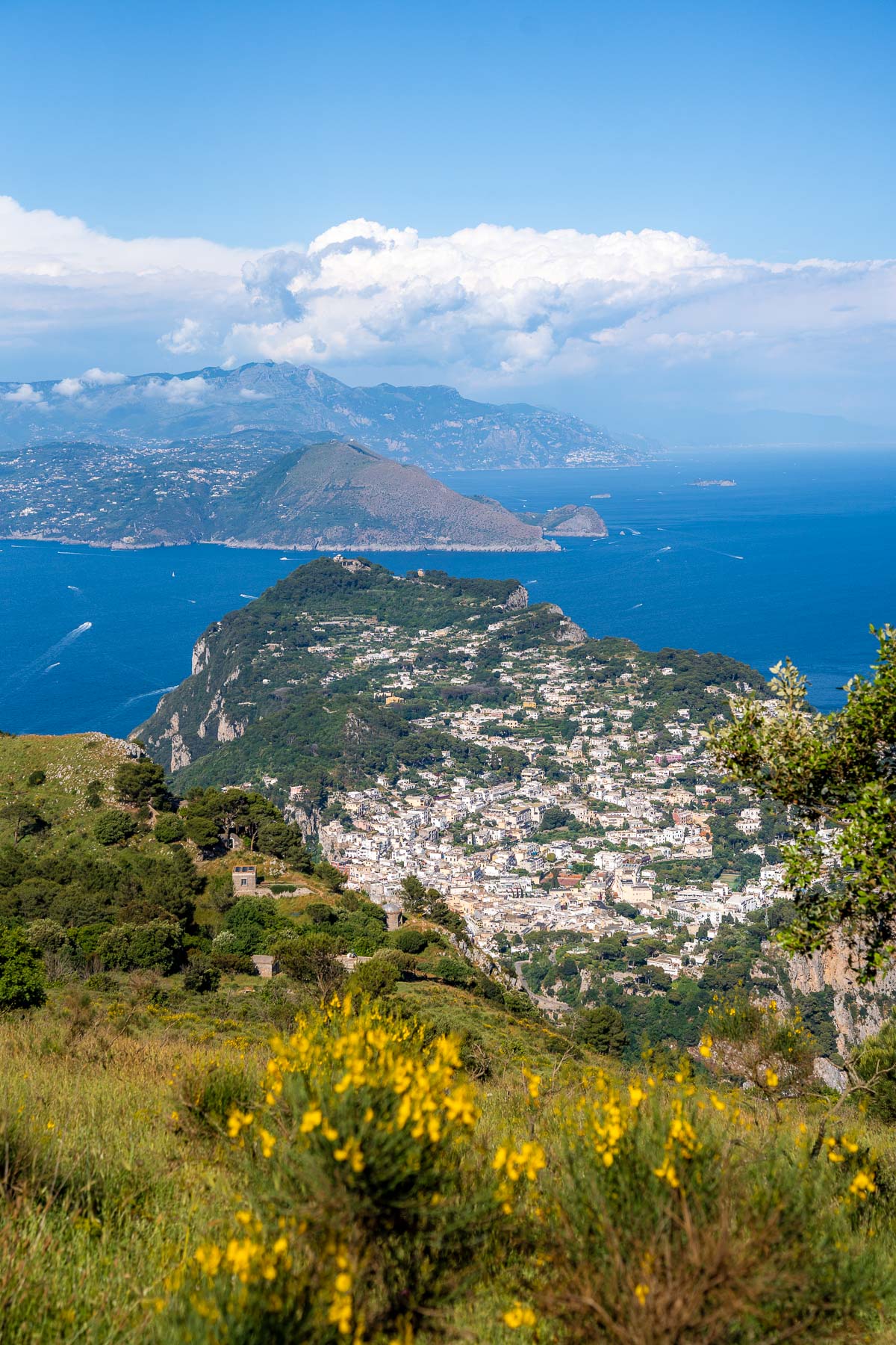 View from Monte Solaro in Capri overlooking the island below, with clustered white buildings, green hills, deep blue sea, and distant coastline under a clear sky.