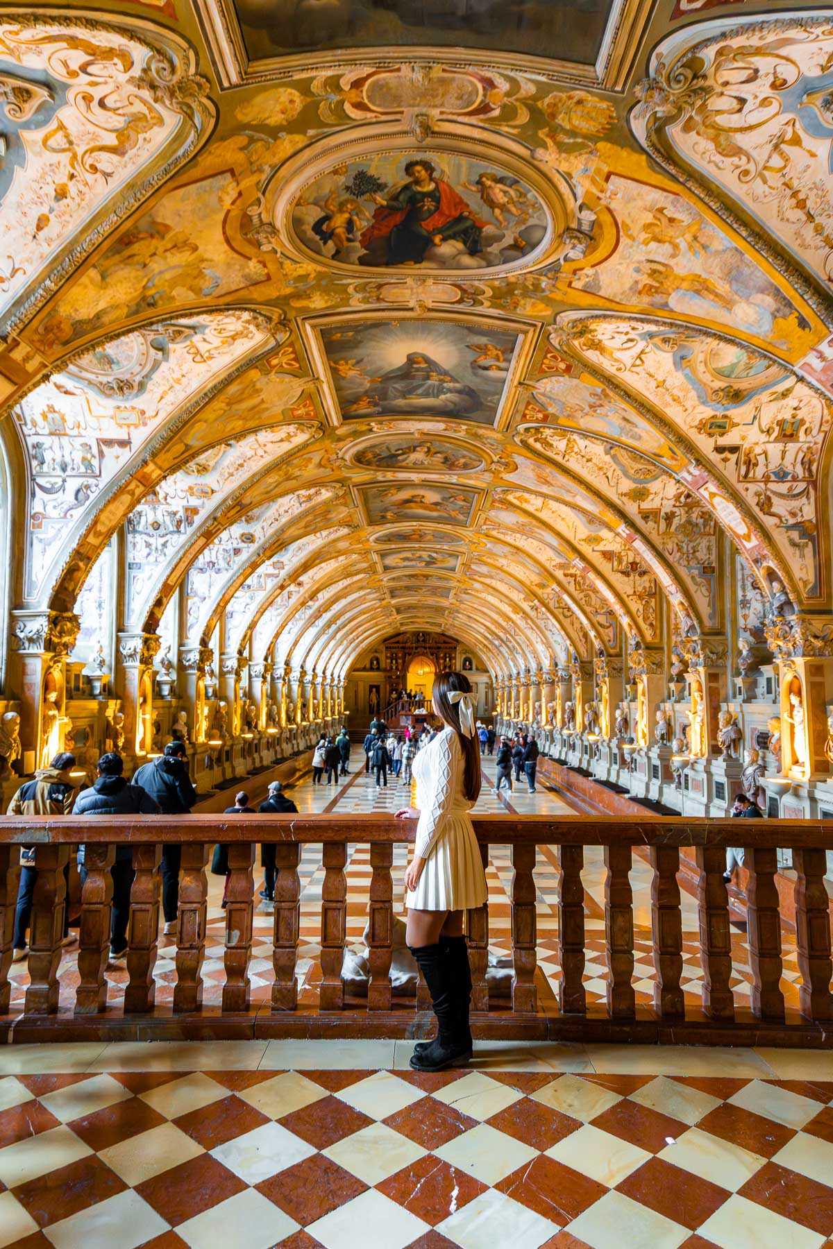A woman stands at a wooden railing inside the Residence Museum in Munich, looking down the long, arched hall lined with statues, warm lights, and an ornate painted ceiling.