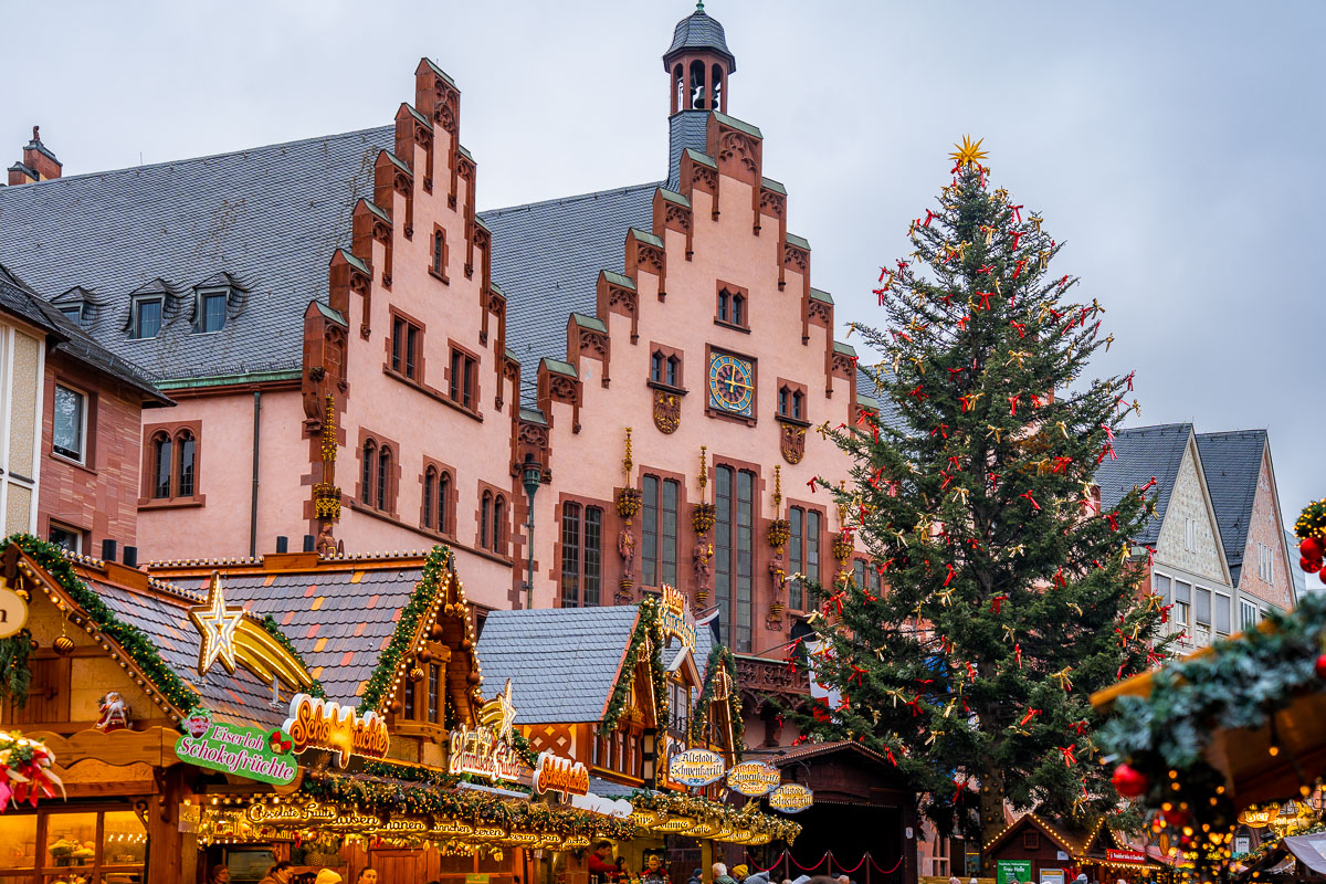Daytime view of the Römerberg Christmas Market, Frankfurt, showing the Römer building behind a tall decorated Christmas tree and wooden market stalls filled with people.