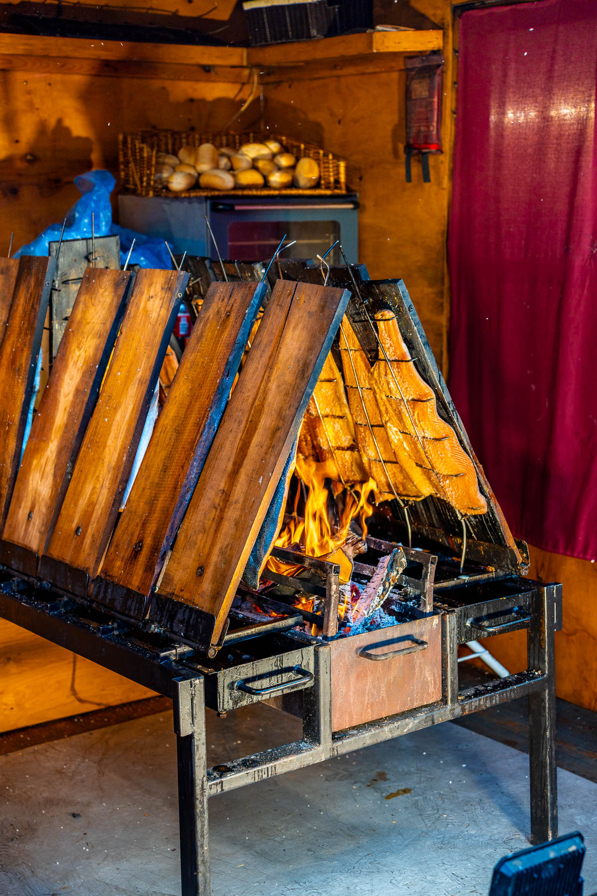 Close-up of salmon grilling over an open fire at Winterwald in Hamburg, with wooden planks leaning over the flames and bread stacked on a shelf in the background.