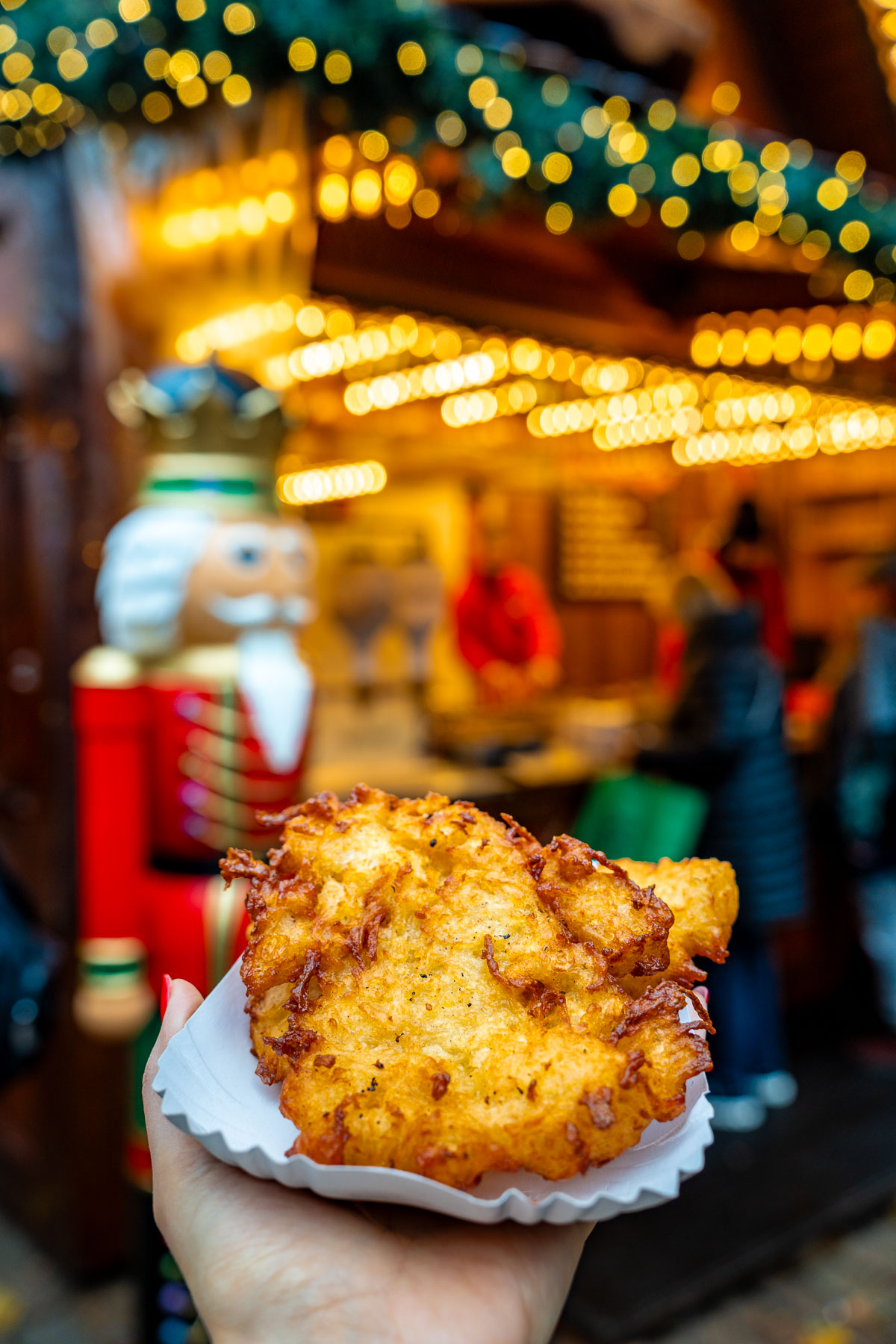 Hand holding a potato pancake at Winterwald in Hamburg, with a nutcracker figure and softly lit market stalls blurred in the background.