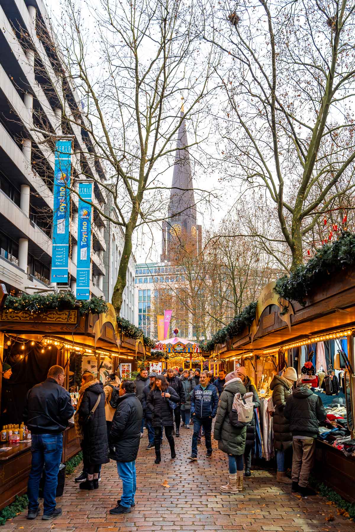 Walkway through Winterwald in Hamburg with wooden market stalls on both sides, people browsing and walking through, and St. Michael’s Church tower visible in the distance.