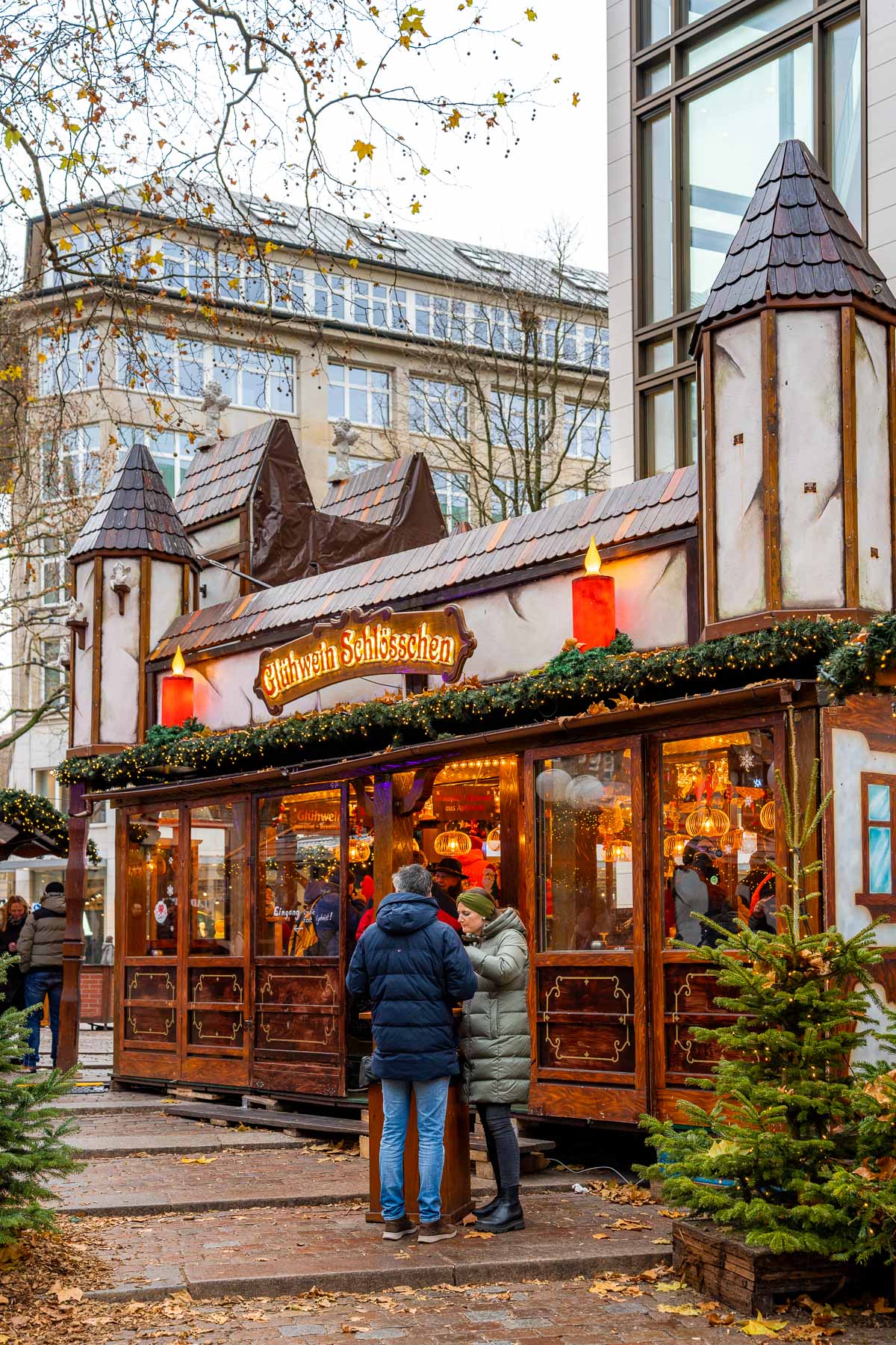 Glühwein stand at Winterwald in Hamburg designed like a small wooden house, with people standing at tall tables, warm lights inside, and small fir trees placed around the stall.