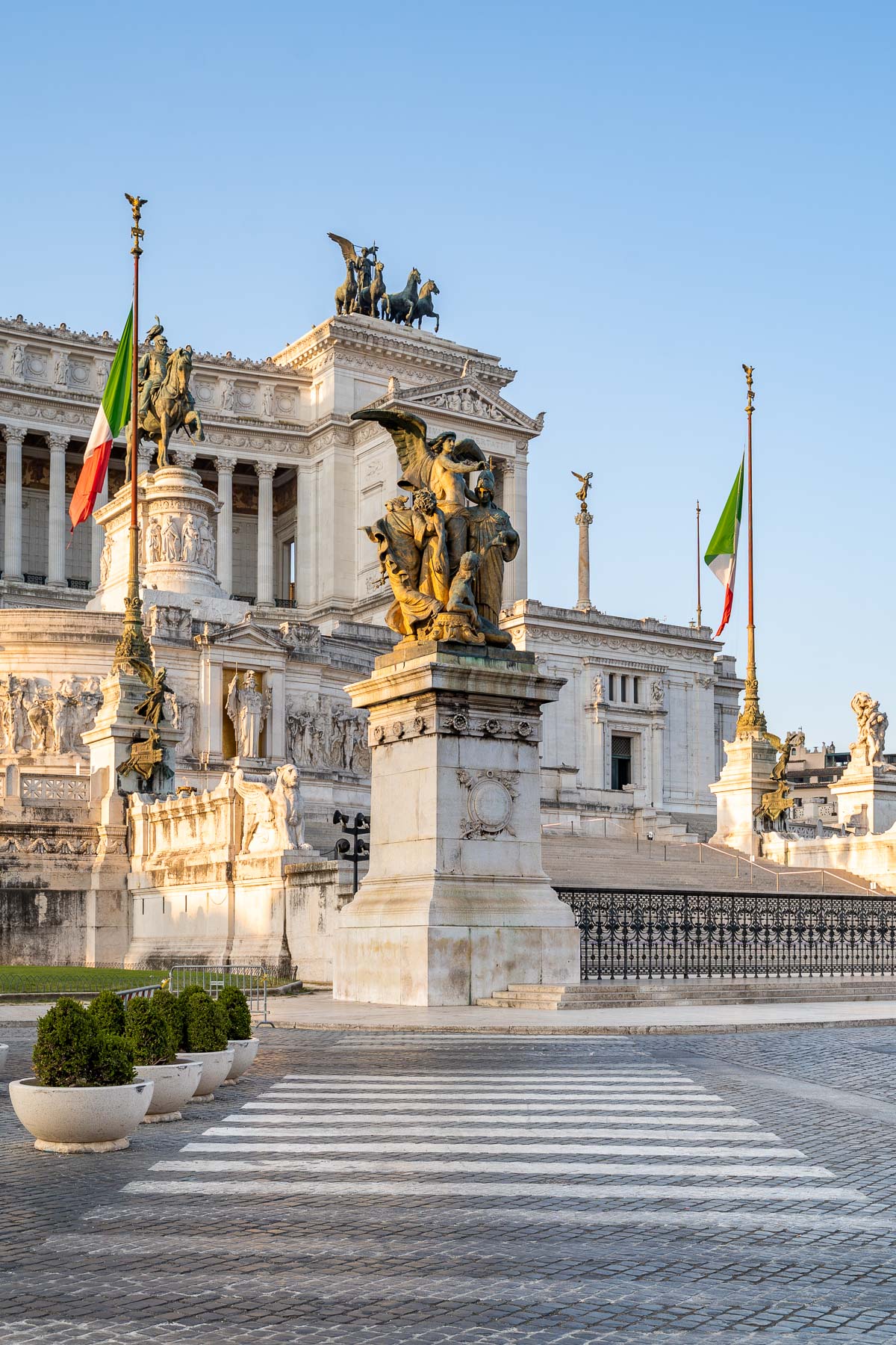 The Altare della Patria in Rome stands in warm evening light, with statues, columns, Italian flags, and a crosswalk leading toward the monument.