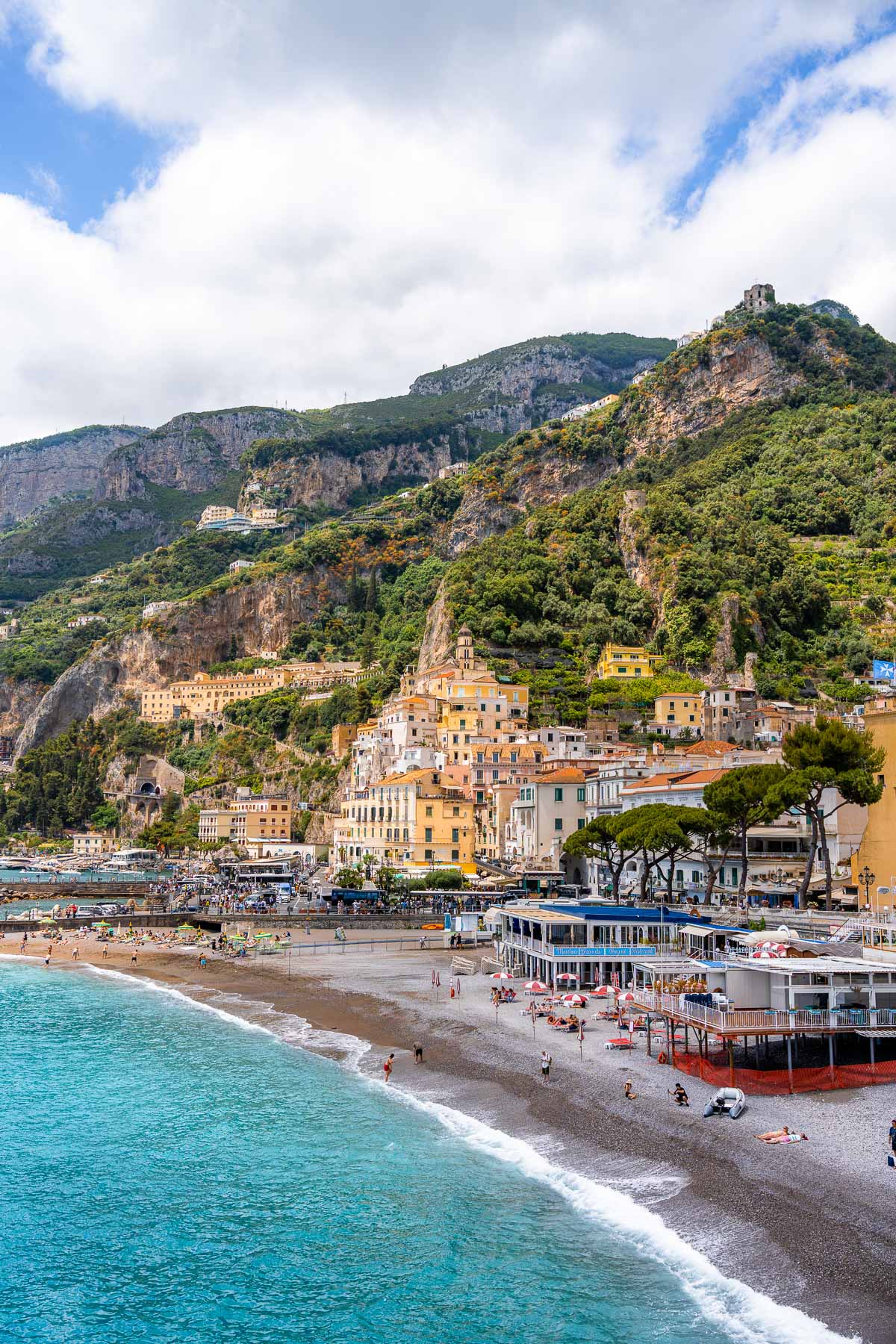A wide view of Amalfi, Italy, with pastel buildings climbing the cliffs above the beach, turquoise water along the shoreline, and beach clubs and umbrellas lining the sand.