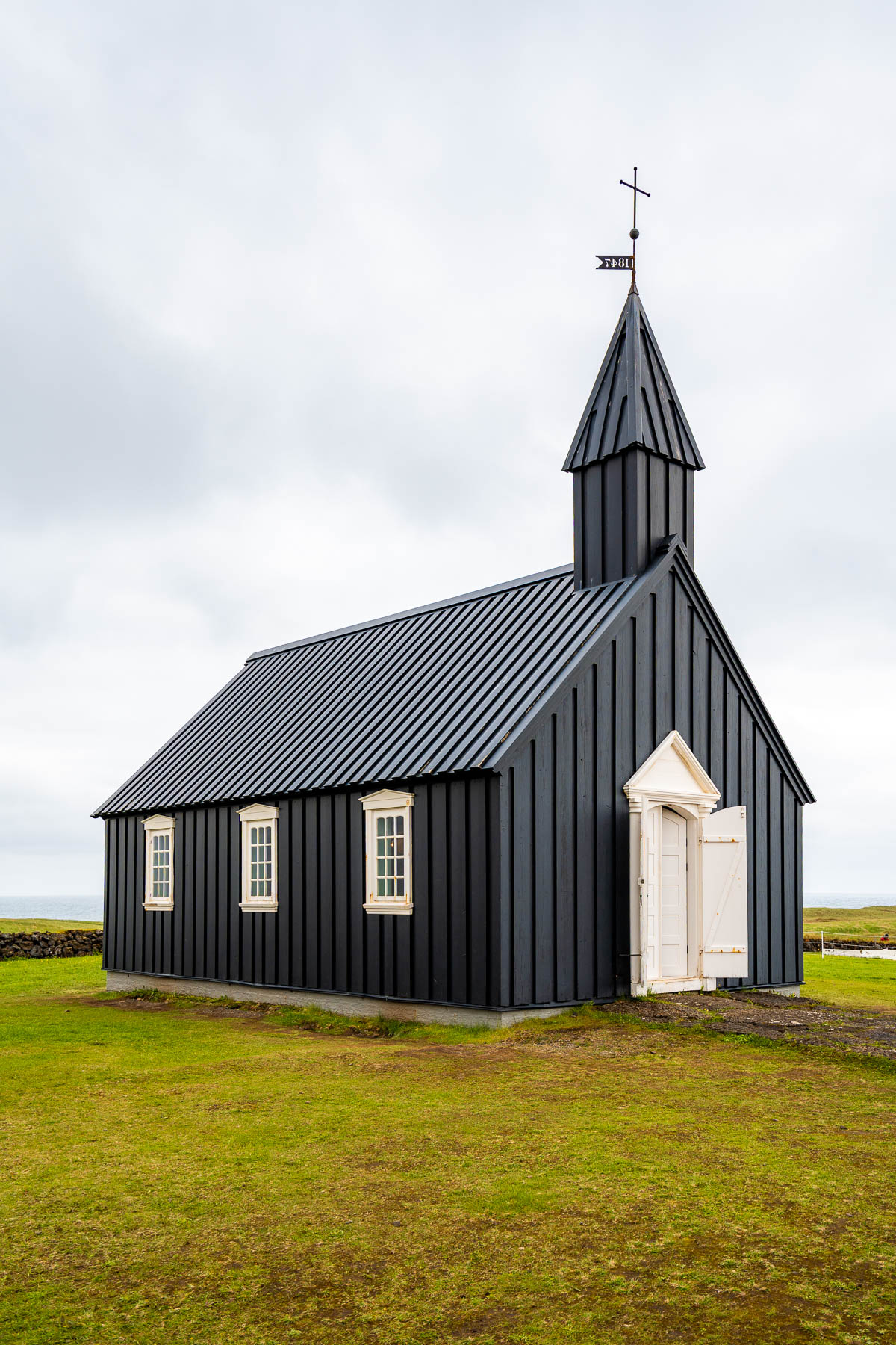 The black wooden church Budakirkja, Iceland sits on a grassy field near the coast with white-framed windows and the ocean in the background.