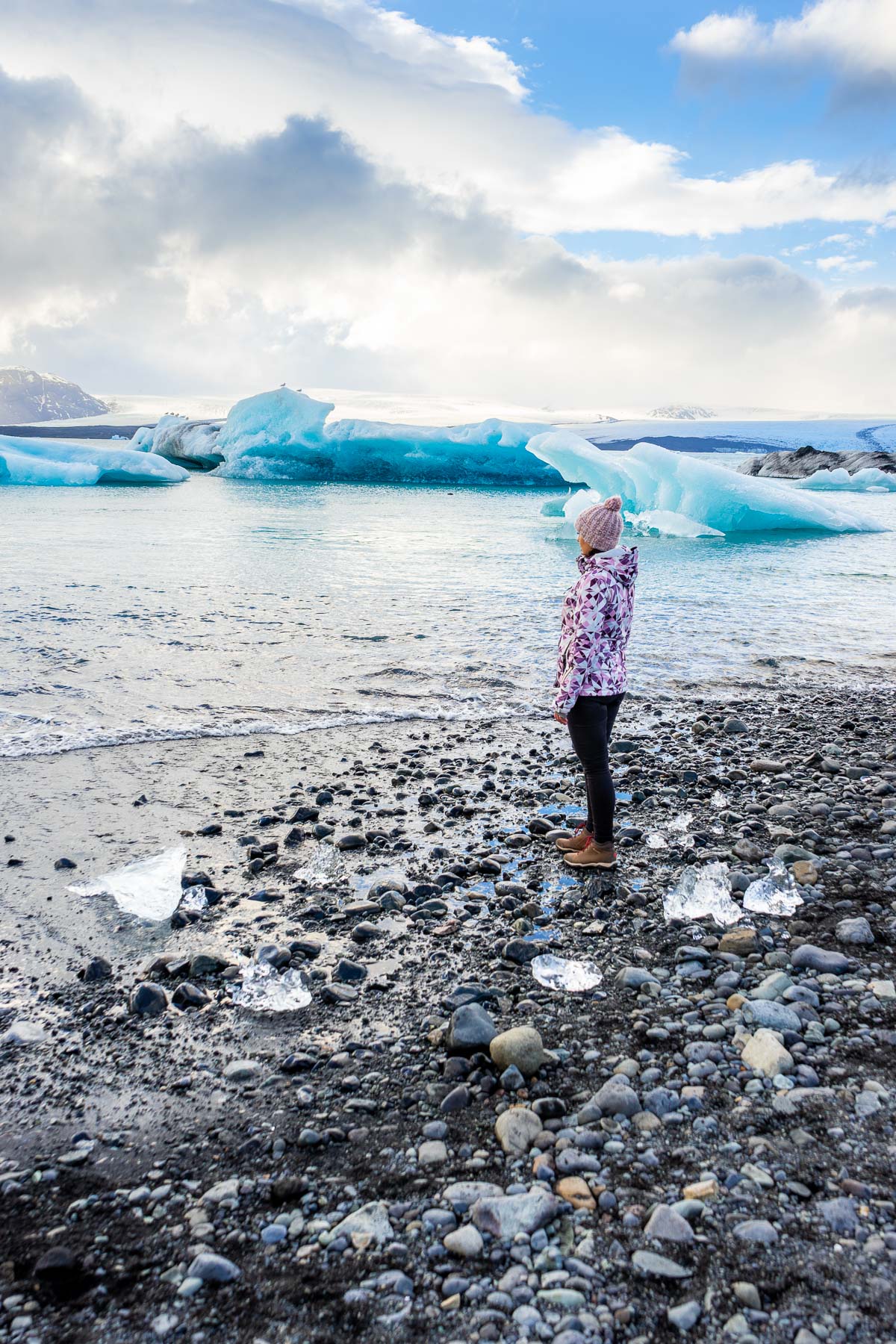 A woman in a pink knit hat and patterned jacket stands on the rocky shore at Diamond Beach, Iceland facing bright blue icebergs in the water.