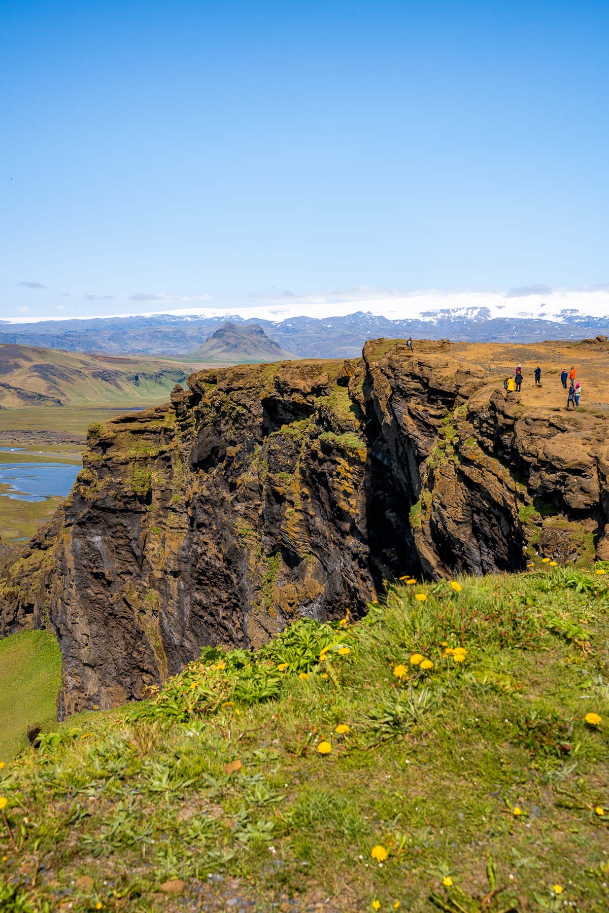 Visitors stand on the cliffs at Dyrholaey, Iceland overlooking dramatic rock formations, green slopes, and distant mountains.