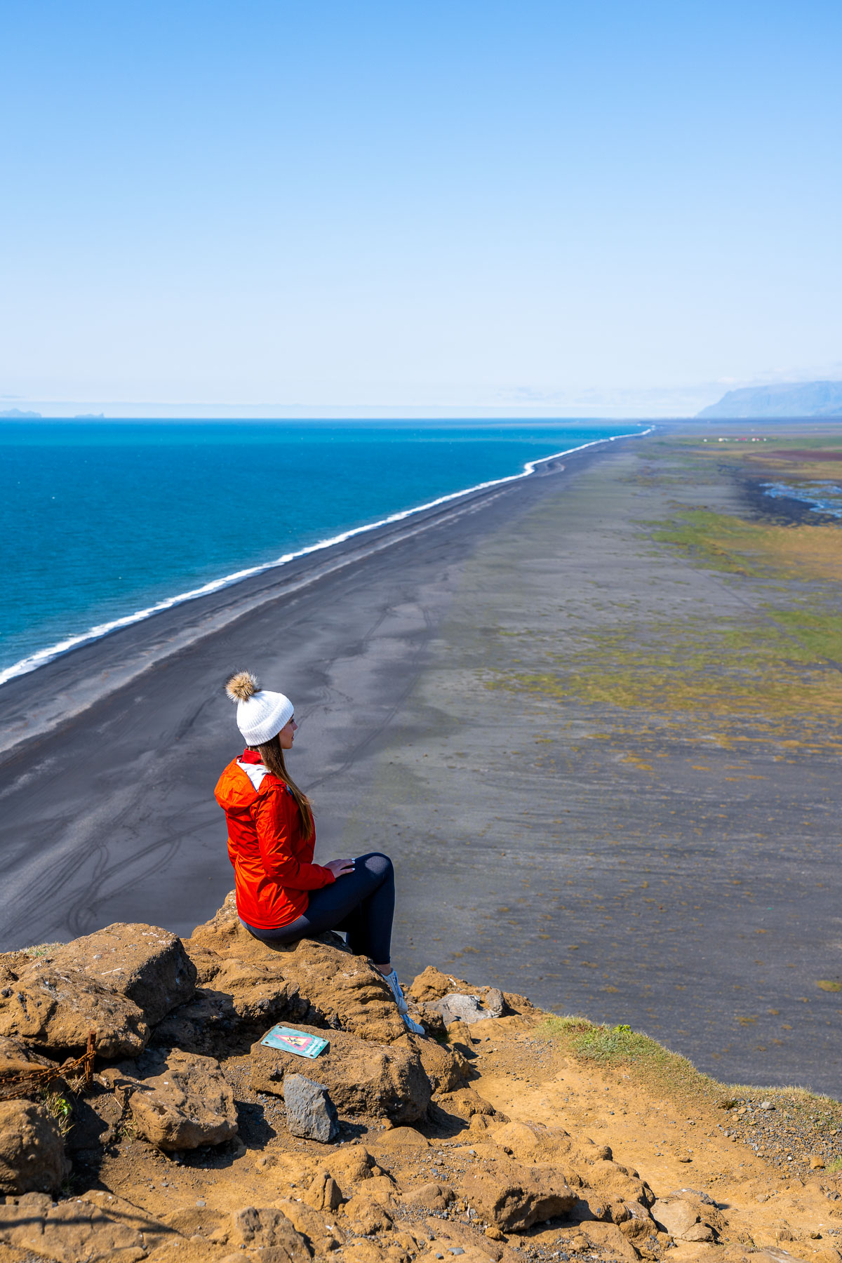 A woman in a red jacket and white beanie sits on a rocky cliff at Dyrholaey, Iceland overlooking the long black sand beach and bright blue ocean.