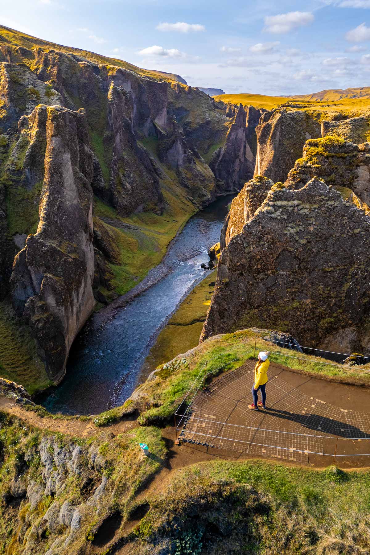 A woman in a yellow jacket stands on a viewing platform at Fjadrargljufur Canyon, Iceland overlooking the deep canyon and river below.