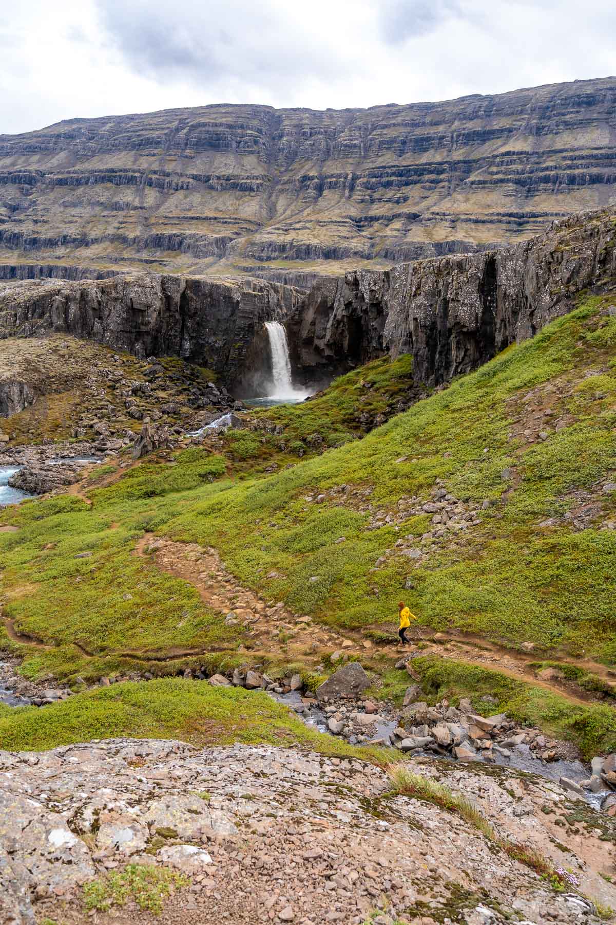 A waterfall flows over a rocky cliff at Folaldafoss, Iceland surrounded by layered mountains and green hills.
