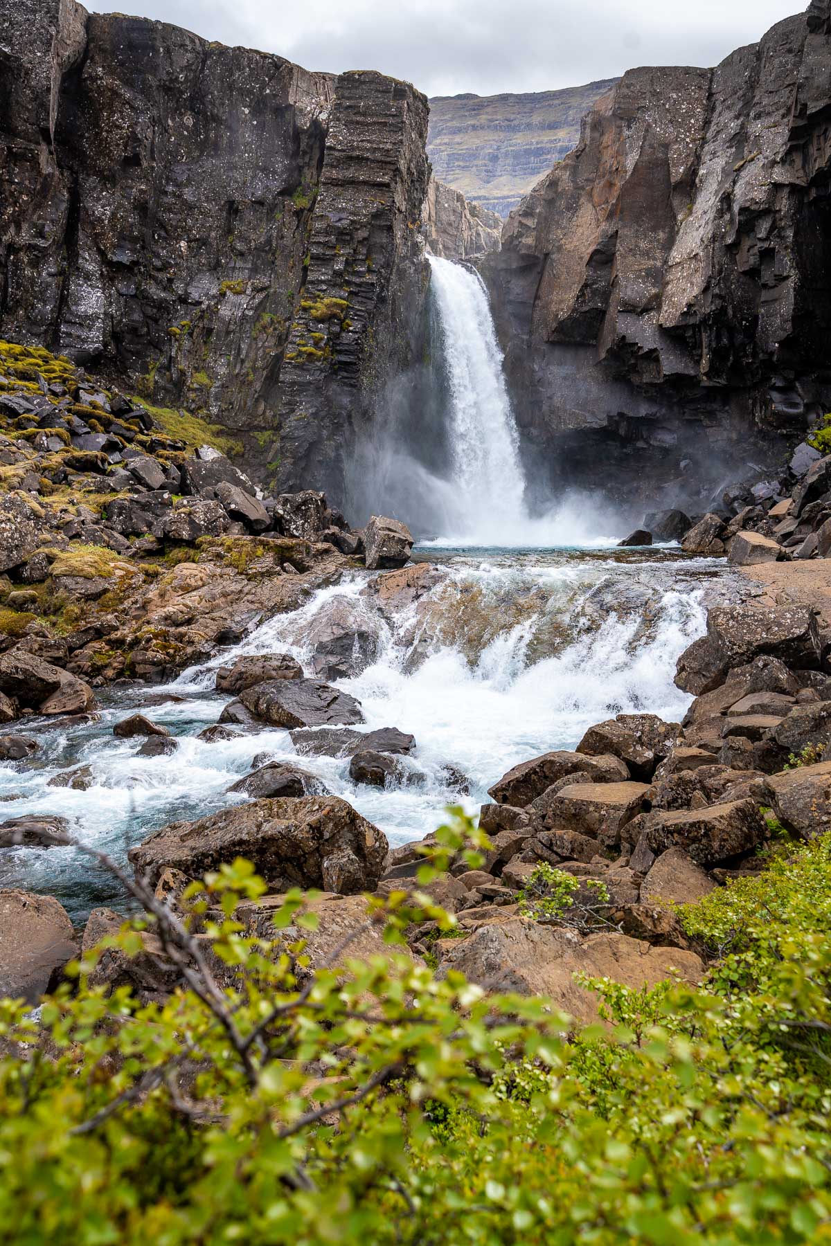 The waterfall at Folaldafoss, Iceland drops between tall dark cliffs with rocky terrain and green shrubs in the foreground.