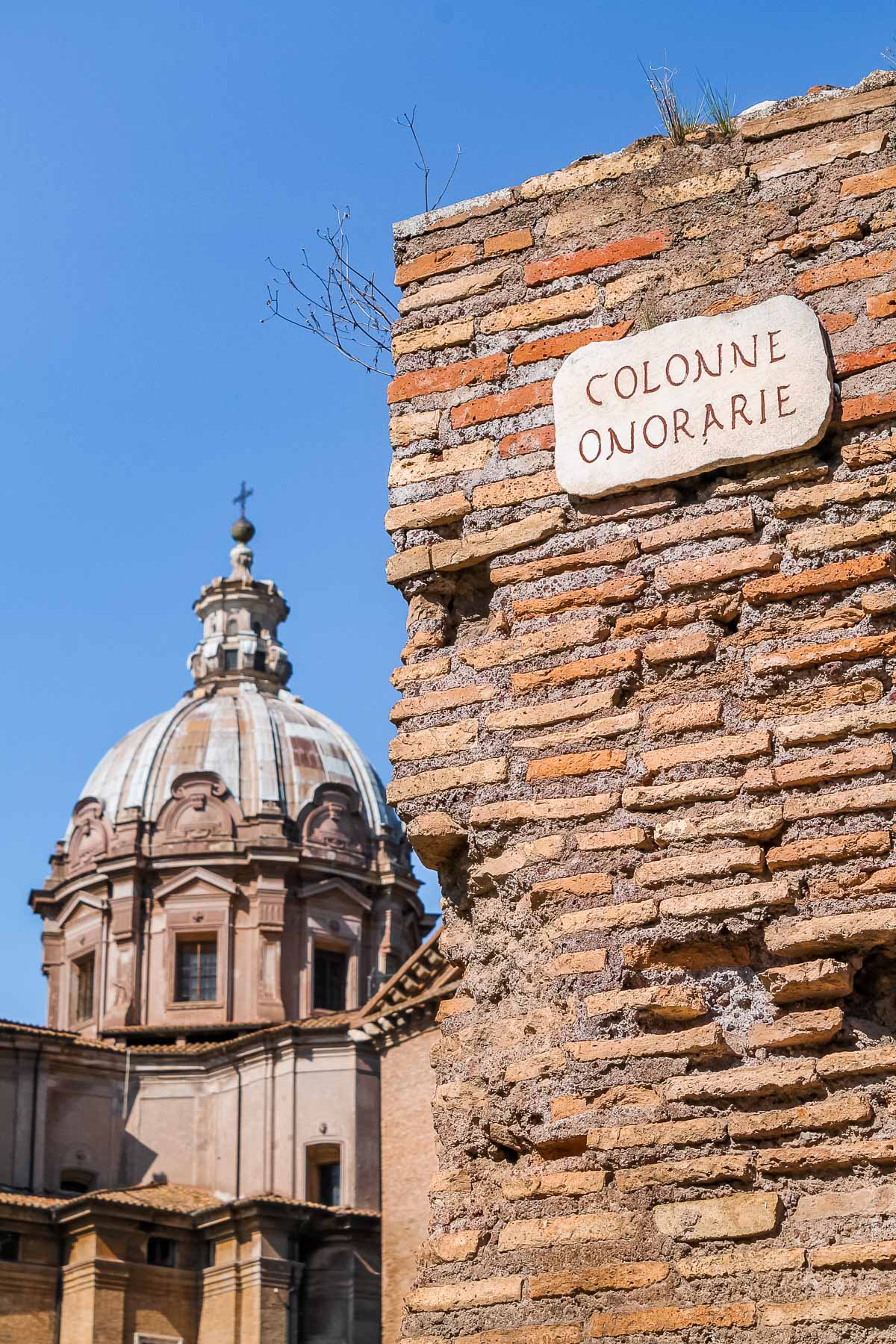 A close-up of an old brick wall in the Forum Romanum in Rome, Italy, with a Colonne Onorarie sign and a domed church visible in the background.