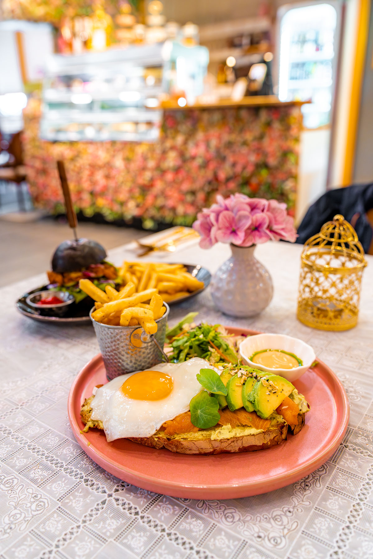 An open-faced sandwich with a fried egg, avocado, and smoked salmon sits on a pink plate at Freyacafe, Iceland with fries and a burger in the background.