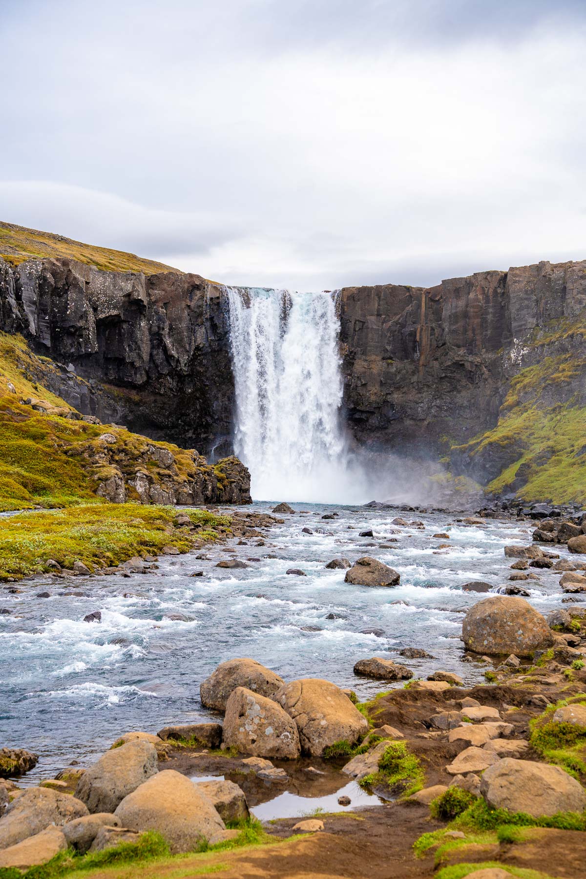 Water from Gufufoss, Iceland crashes into a rocky river below, framed by steep basalt cliffs and green moss.